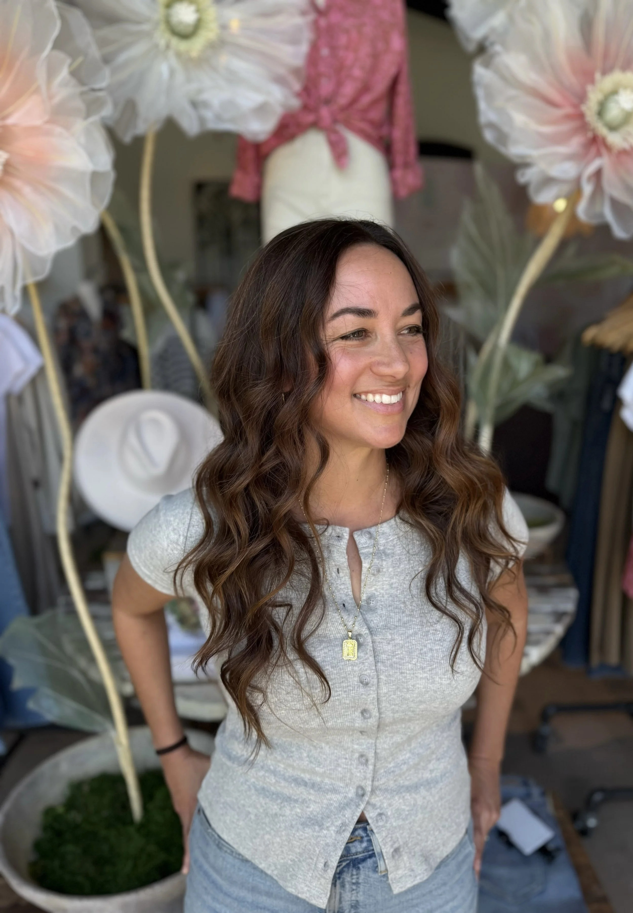 A woman with long, wavy brown hair smiling, standing in front of large decorative flowers and hats.