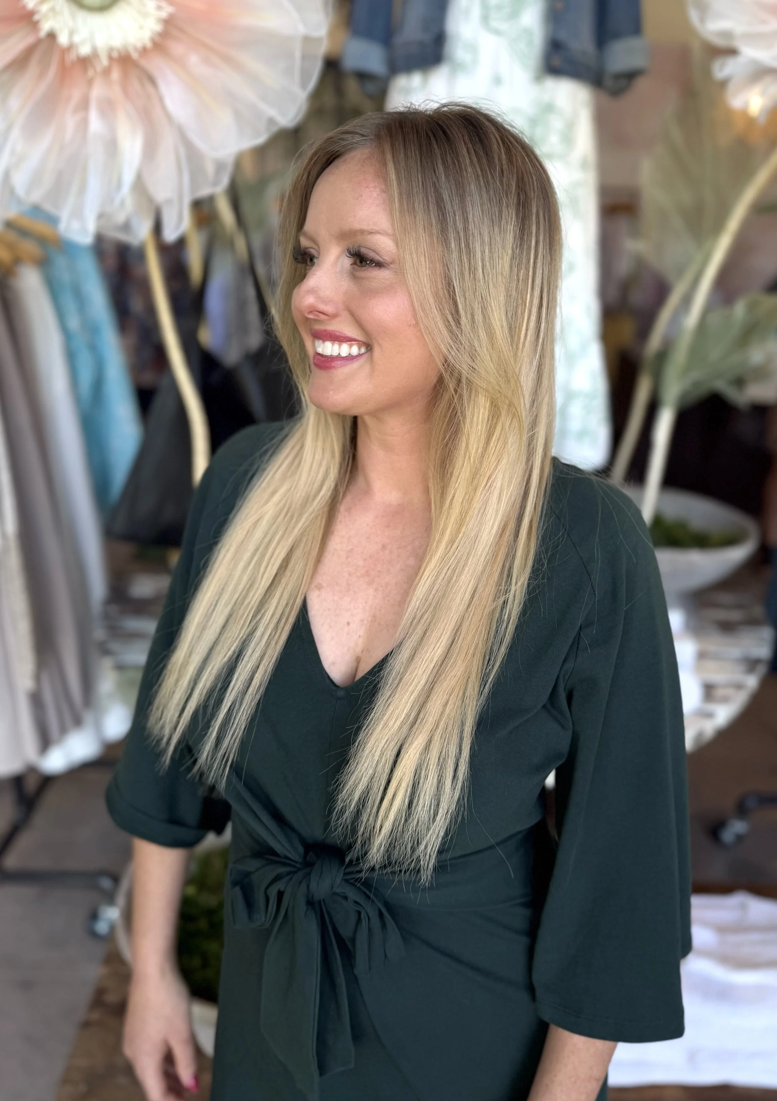 A woman with long blonde hair, smiling and looking to the side, wearing a black dress with a tied waist, standing in a boutique with clothing and decorative umbrellas in the background.