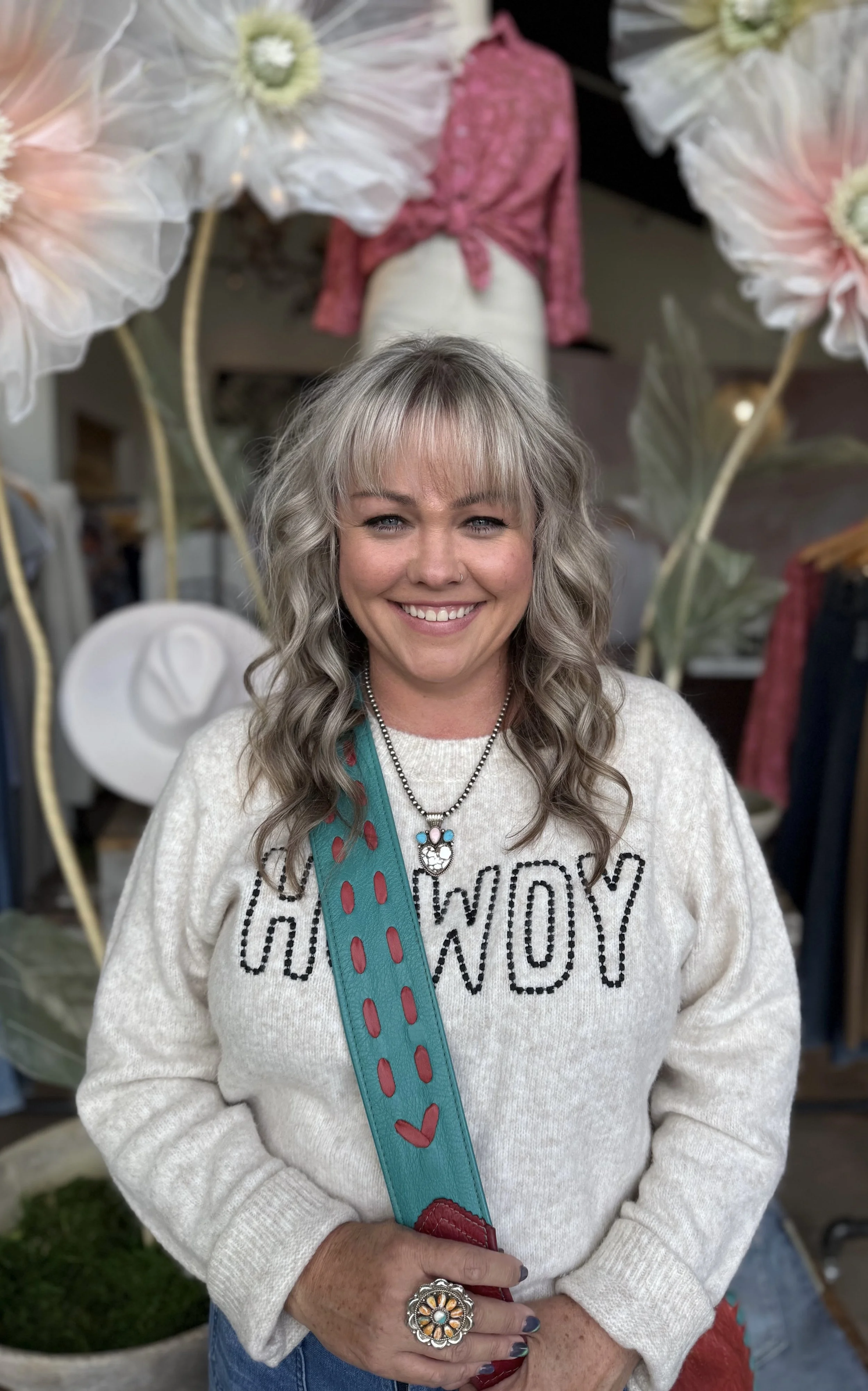 Smiling woman with wavy blonde hair, wearing a white sweater with black lettering, a necklace, and a large ring, standing in front of large flower decorations with a pink blouse mannequin in the background.