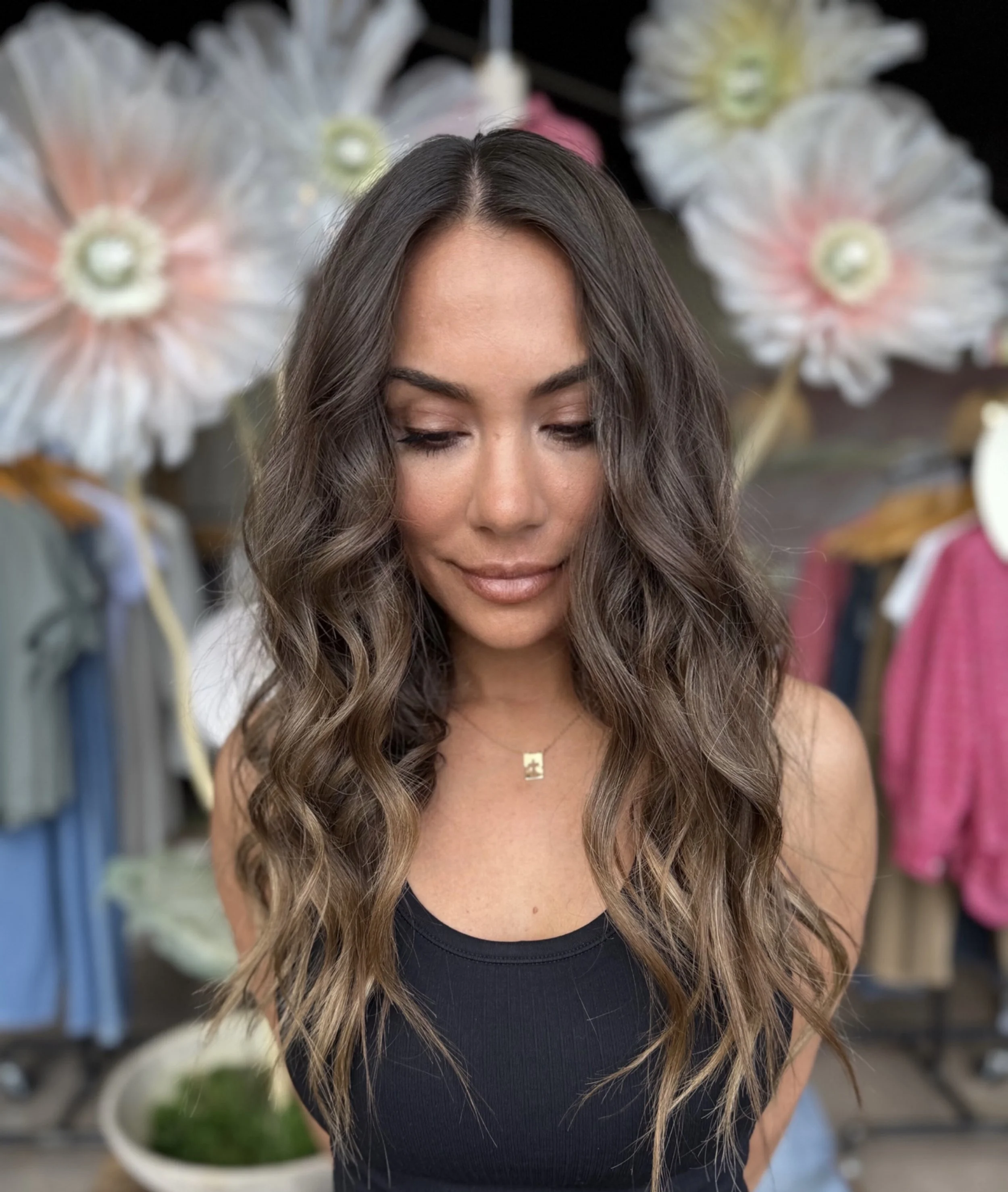 A woman with long wavy brown hair and a tan complexion, wearing a black sleeveless top and a small gold necklace, stands in front of large floral decorations and clothing racks.
