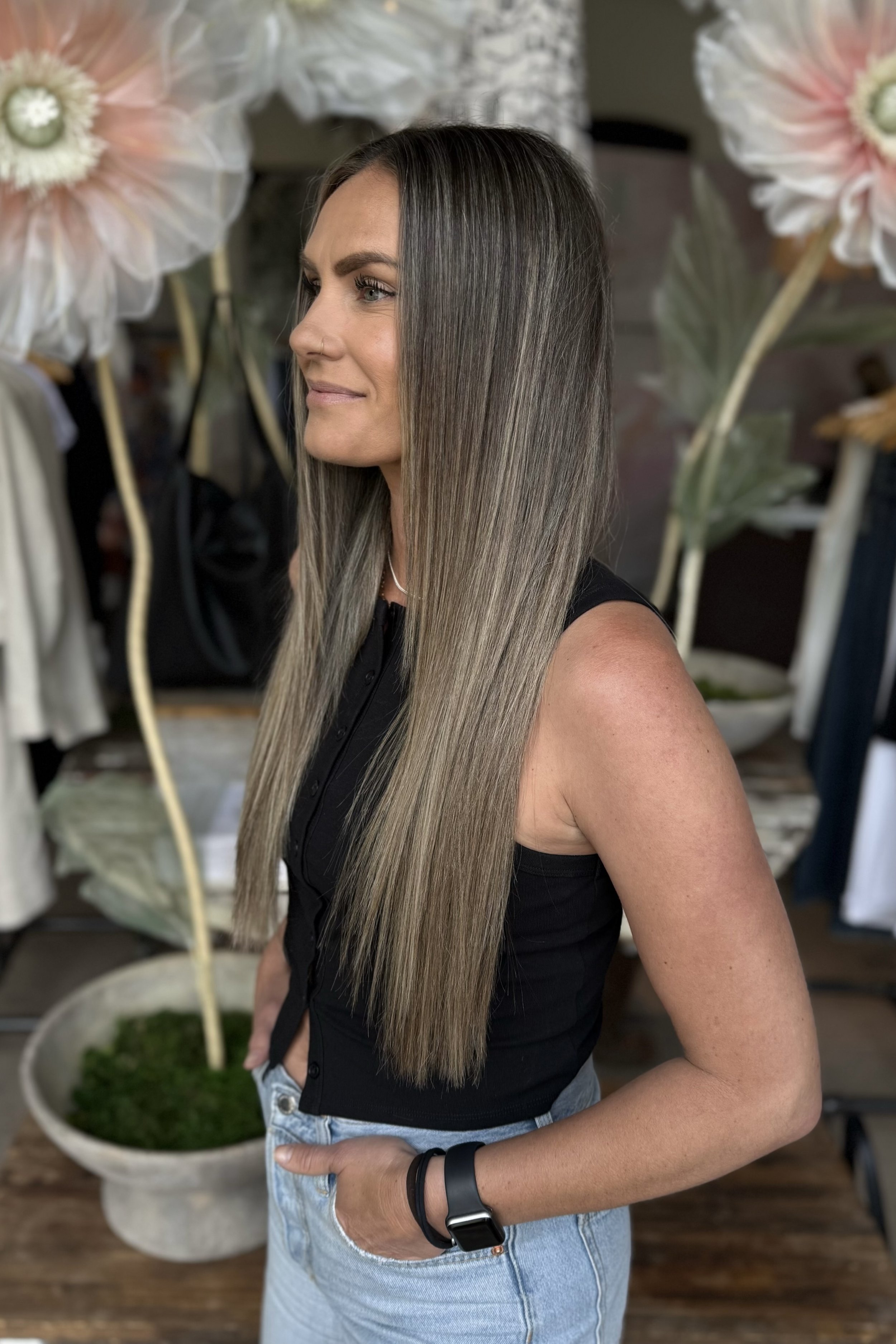 A woman with straight, shoulder-length hair that is dark at the roots and lightens toward the ends, standing indoors with floral decorations in the background.