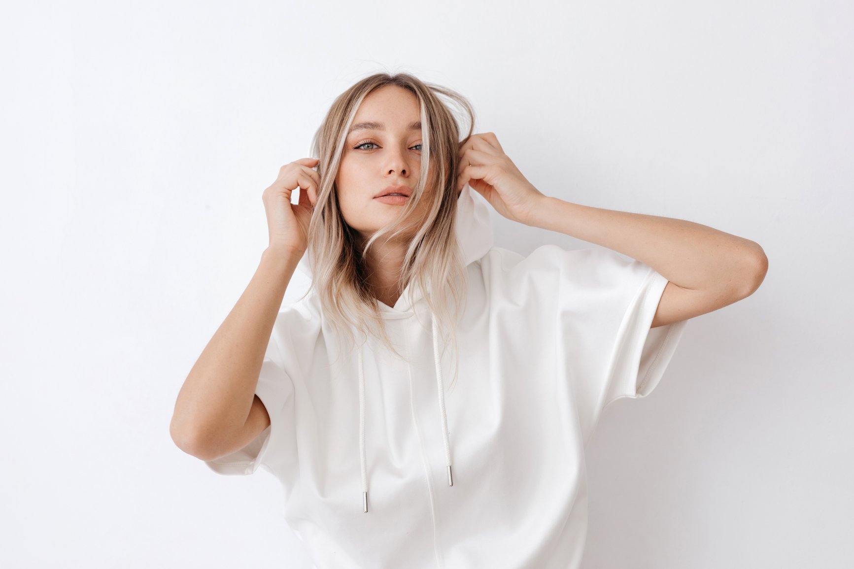 A woman with blonde hair wearing a white hoodie, holding her hair with both hands, standing against a plain white background.