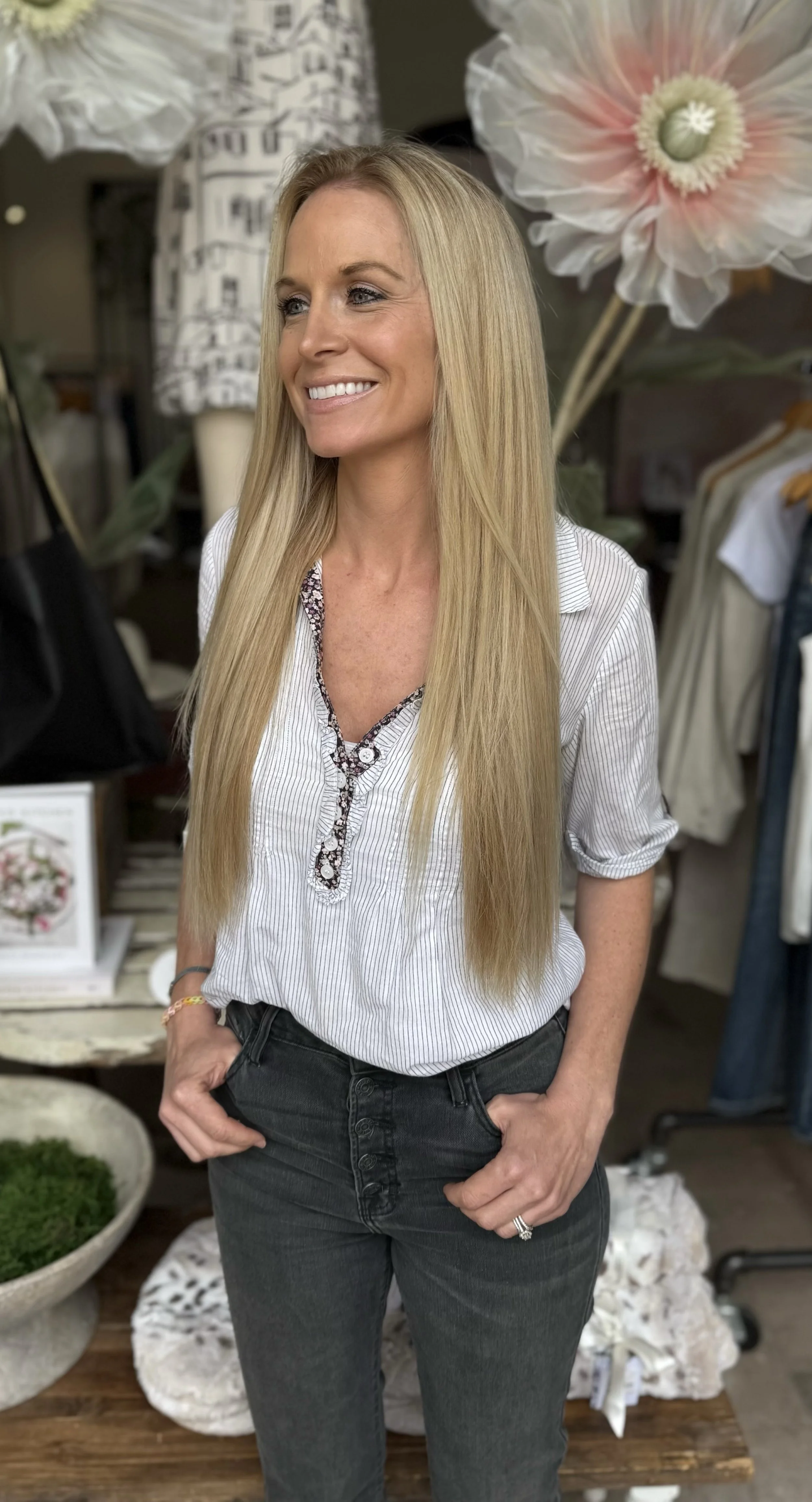 A woman with long blond hair wearing a striped blouse and dark jeans, smiling in a store with large floral decorations and clothing in the background.