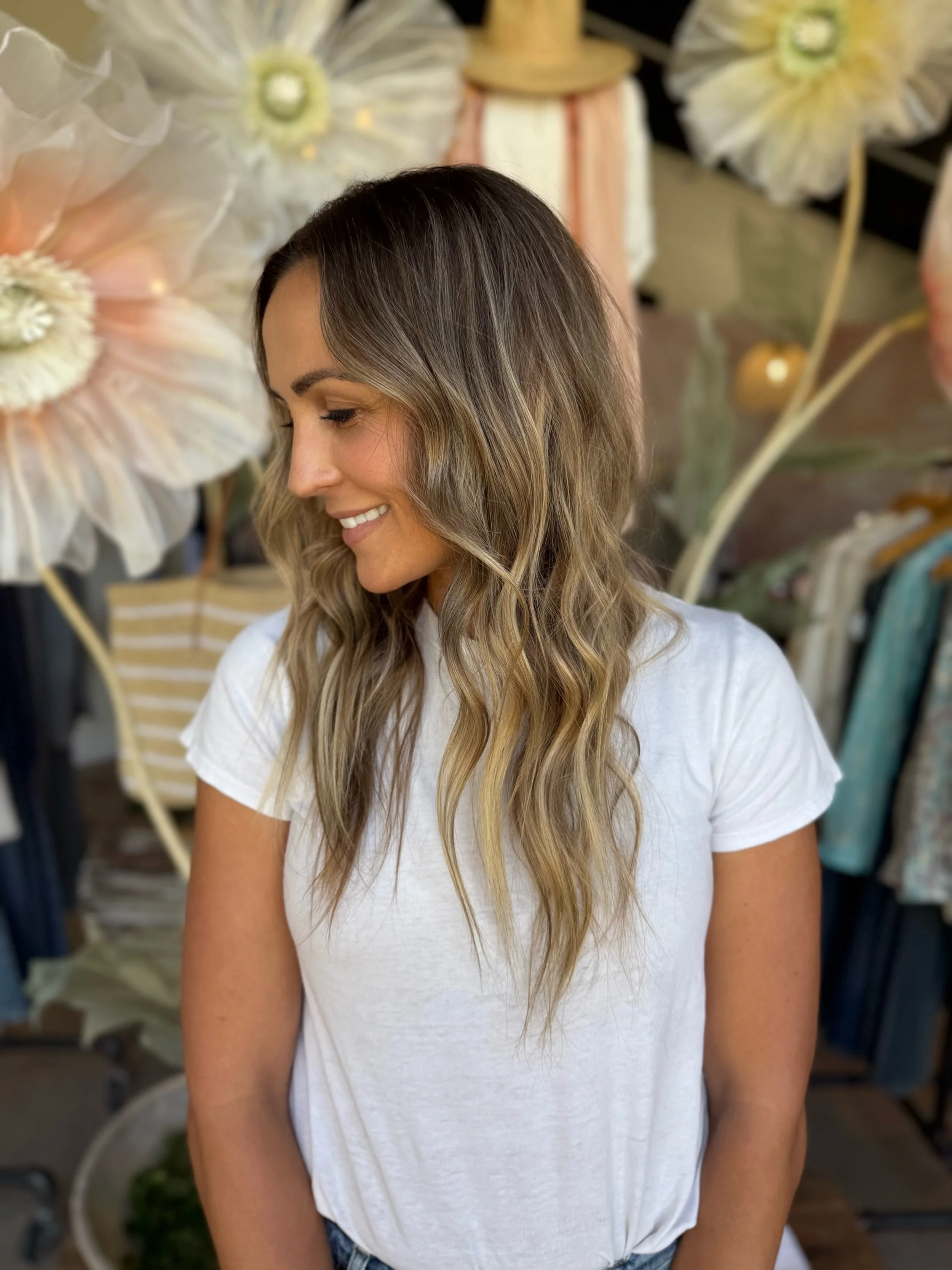 A woman with wavy, shoulder-length hair smiling while looking down, standing in front of large decorative flowers.