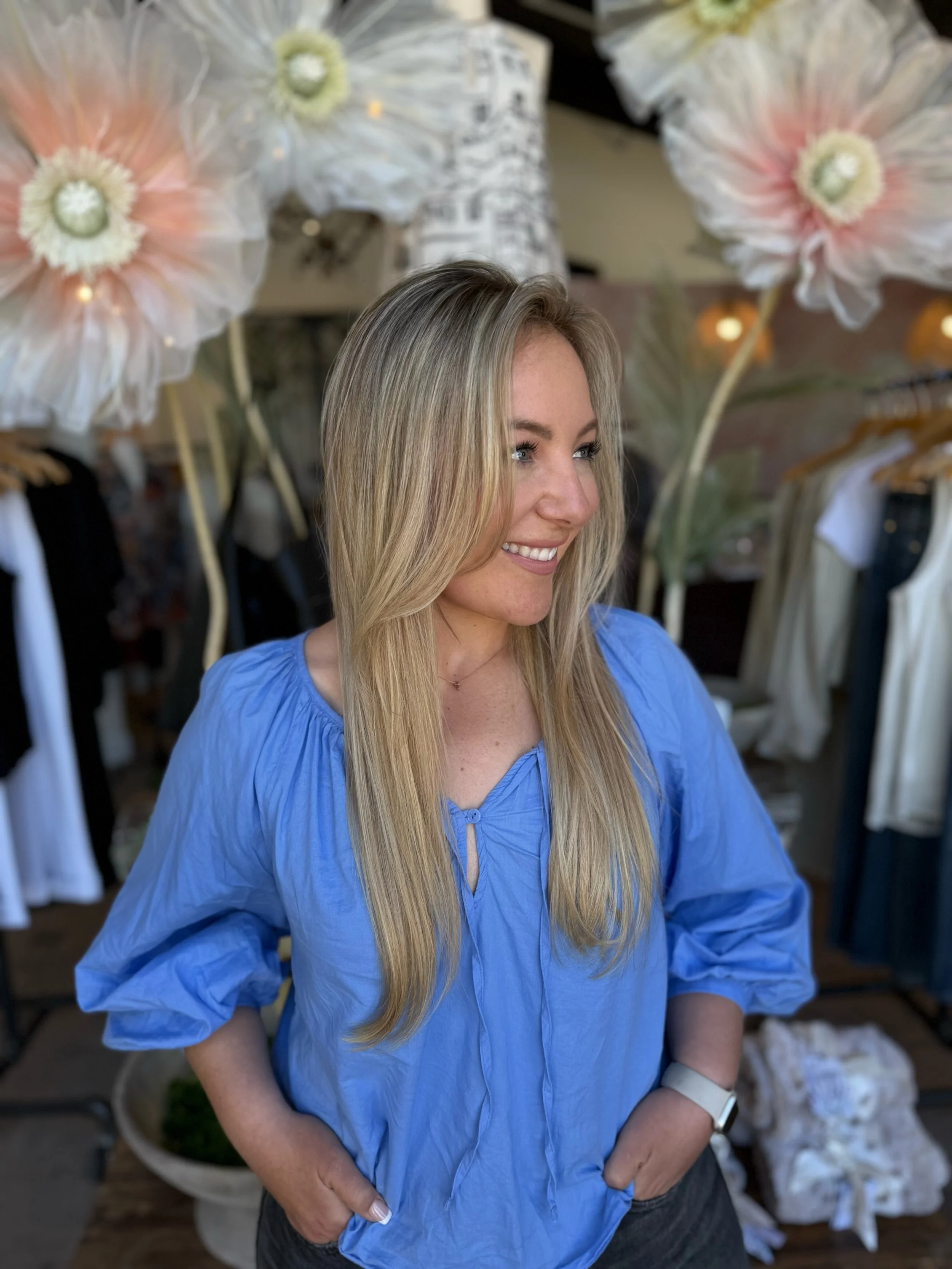 A smiling woman with long blonde hair wearing a blue blouse standing in front of large, decorative paper flowers and clothing racks.