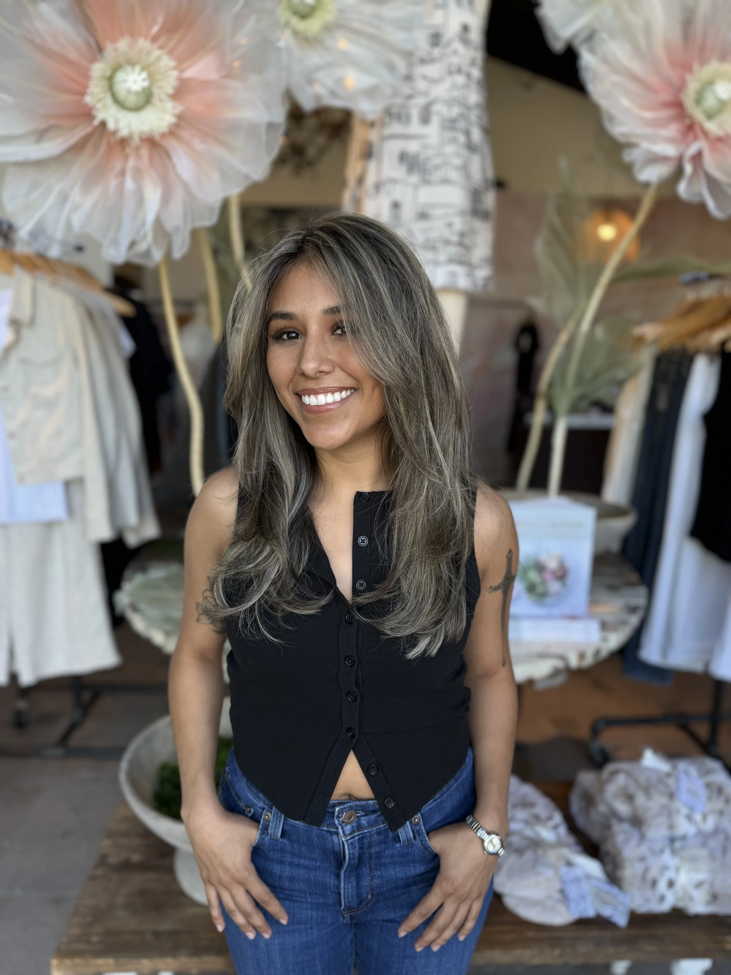 Smiling woman with long brown hair wearing a sleeveless black top and blue jeans, standing in front of a display of large paper flowers and clothing racks.