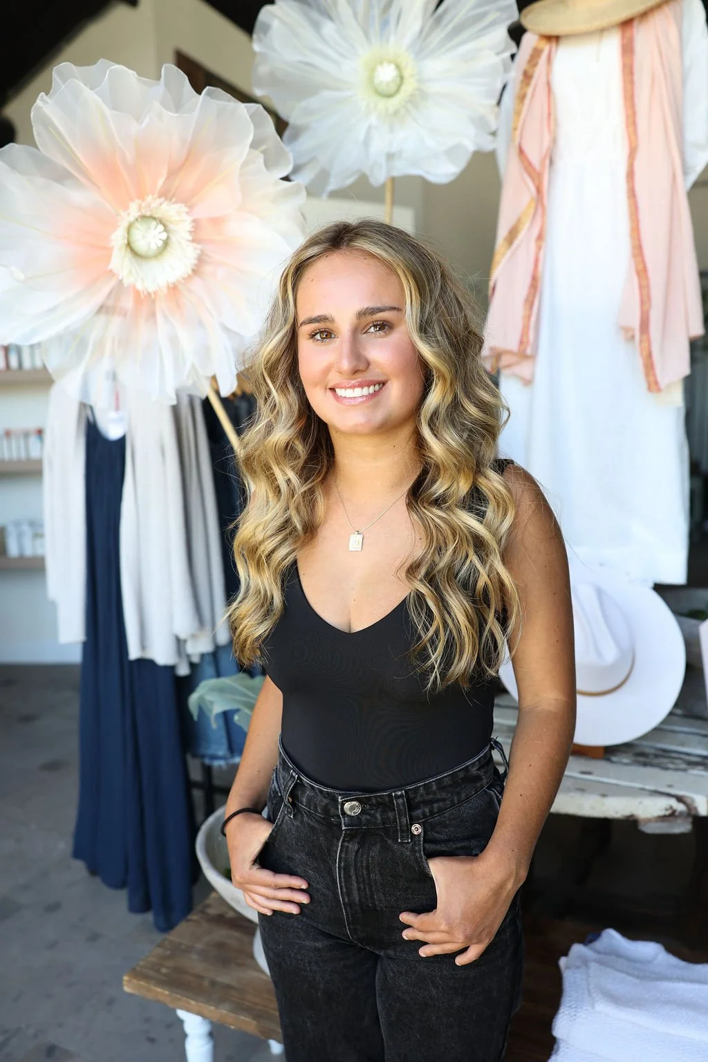 A smiling woman with long, wavy blonde hair, wearing a black tank top and dark jeans, standing indoors with large flower-shaped decorations and clothing in the background.