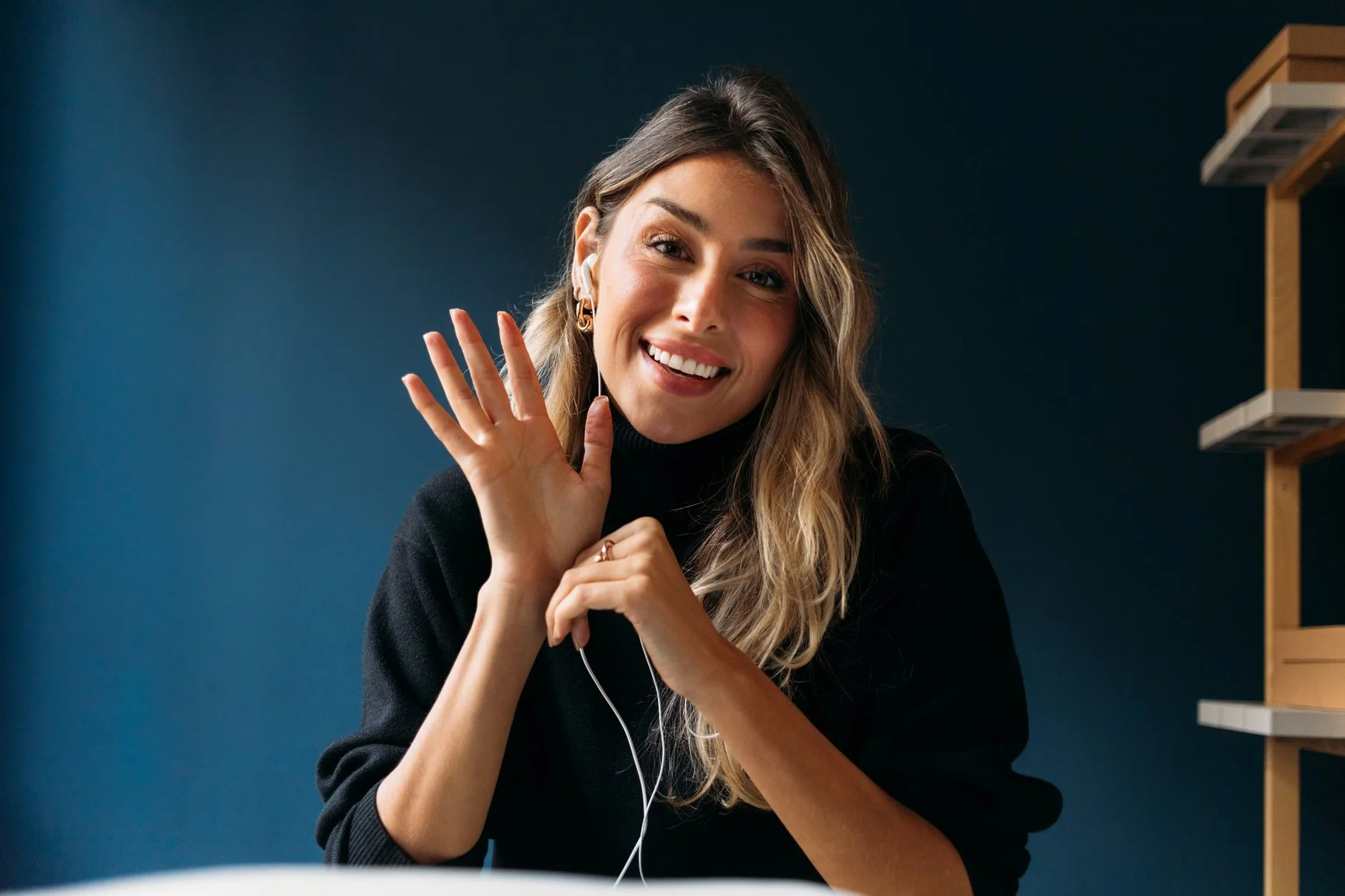 A smiling woman with long wavy hair, wearing a black turtleneck, waving and listening to headphones, in a room with dark blue wall and wooden shelves.