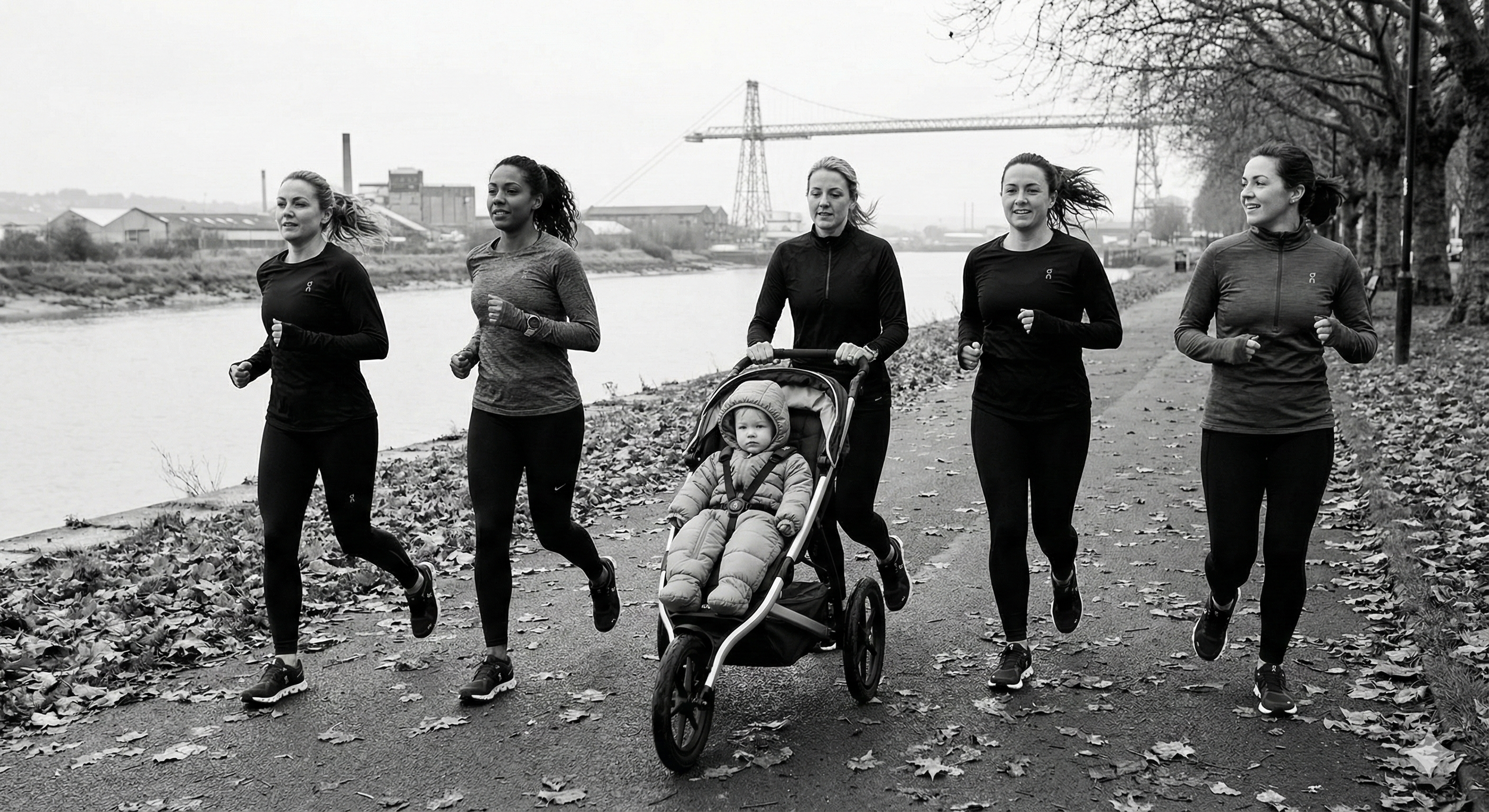 Five women jogging along a riverside path with fallen leaves; one woman is pushing a baby in a stroller. The scene is outdoors with industrial buildings and power lines in the background.