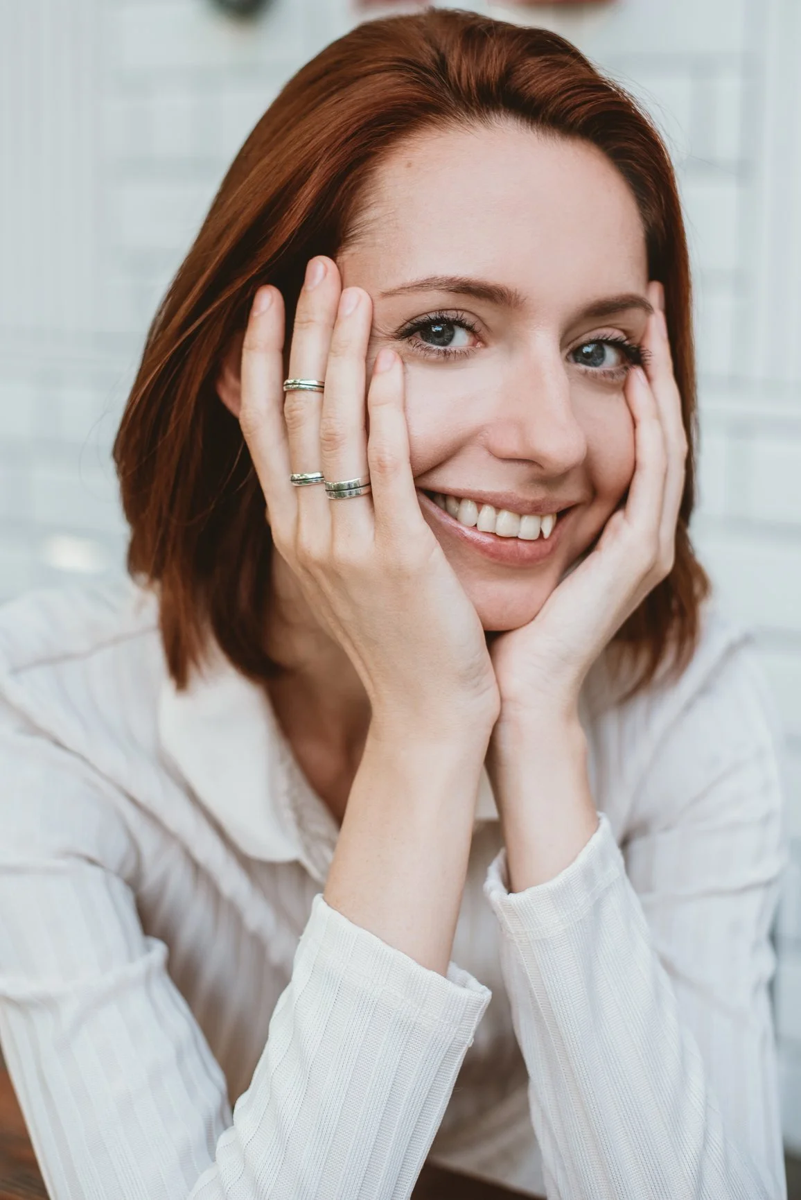 A woman with sleek, red hair smiling while holding her face with her hands, looking calm and happy.