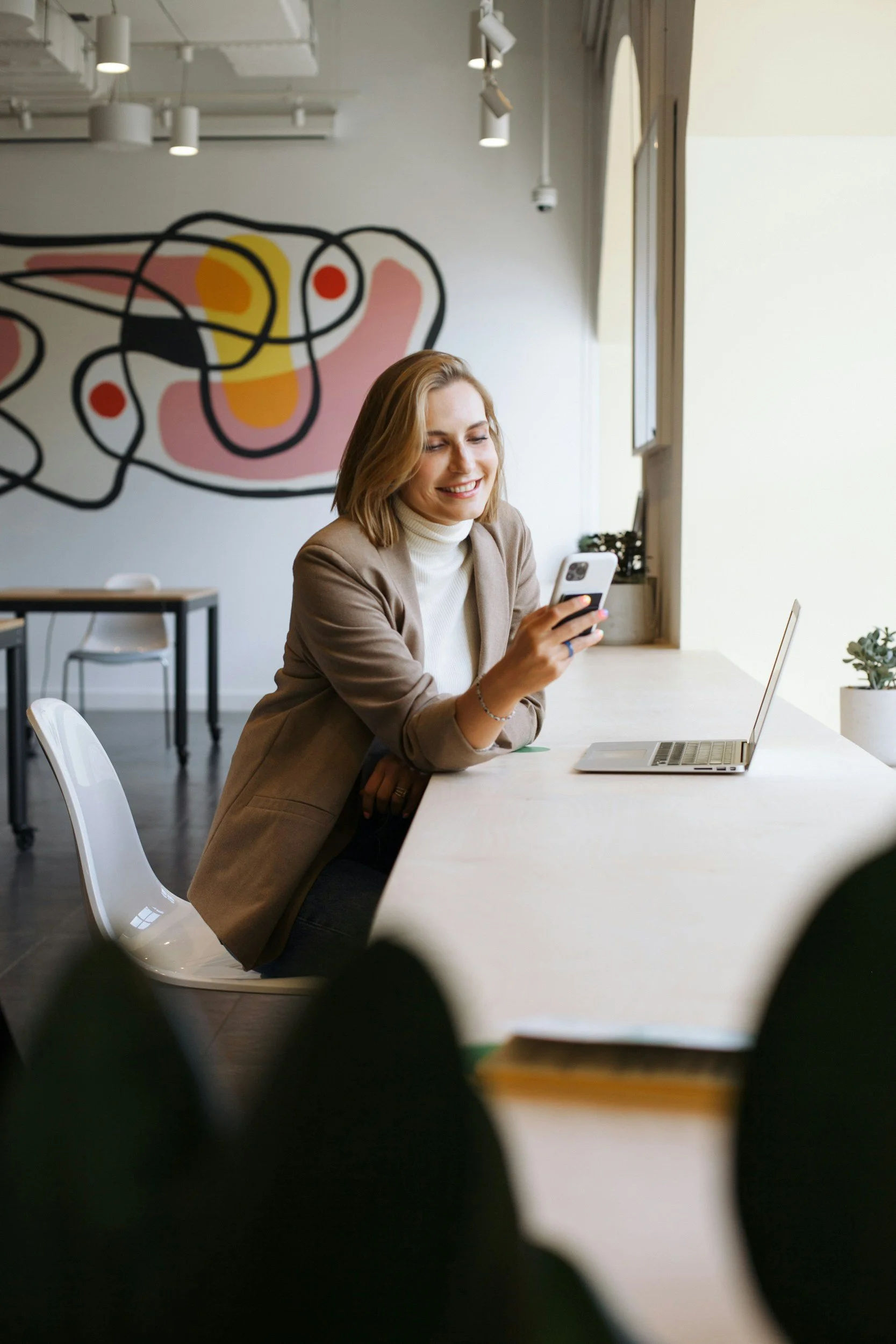 A pretty blonde woman with a bob haircut, smiling at her phone, with a laptop open in front of her looking happy.