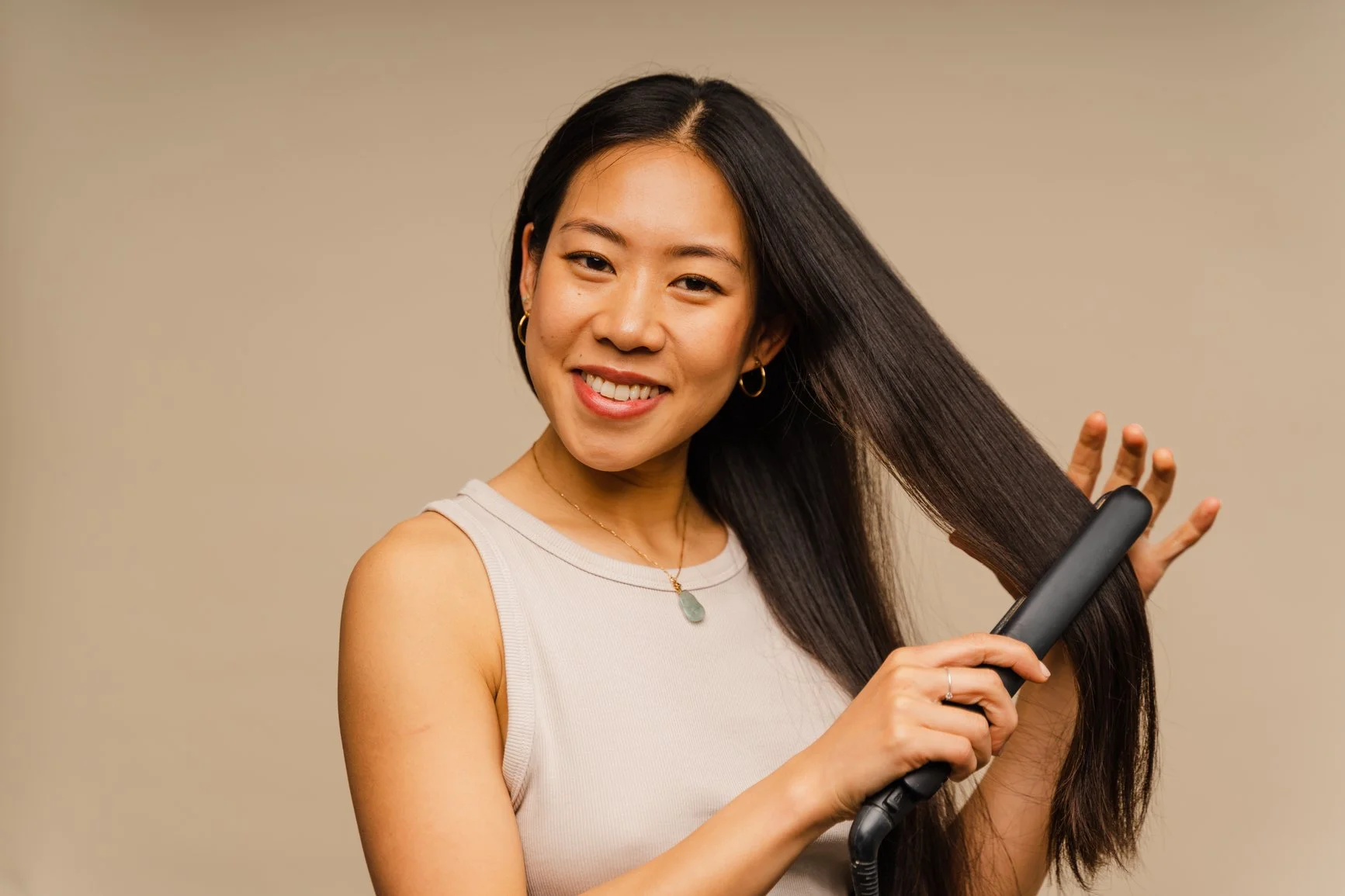 A woman with long dark brown hair smiling and using a flat iron to straighten her hair.