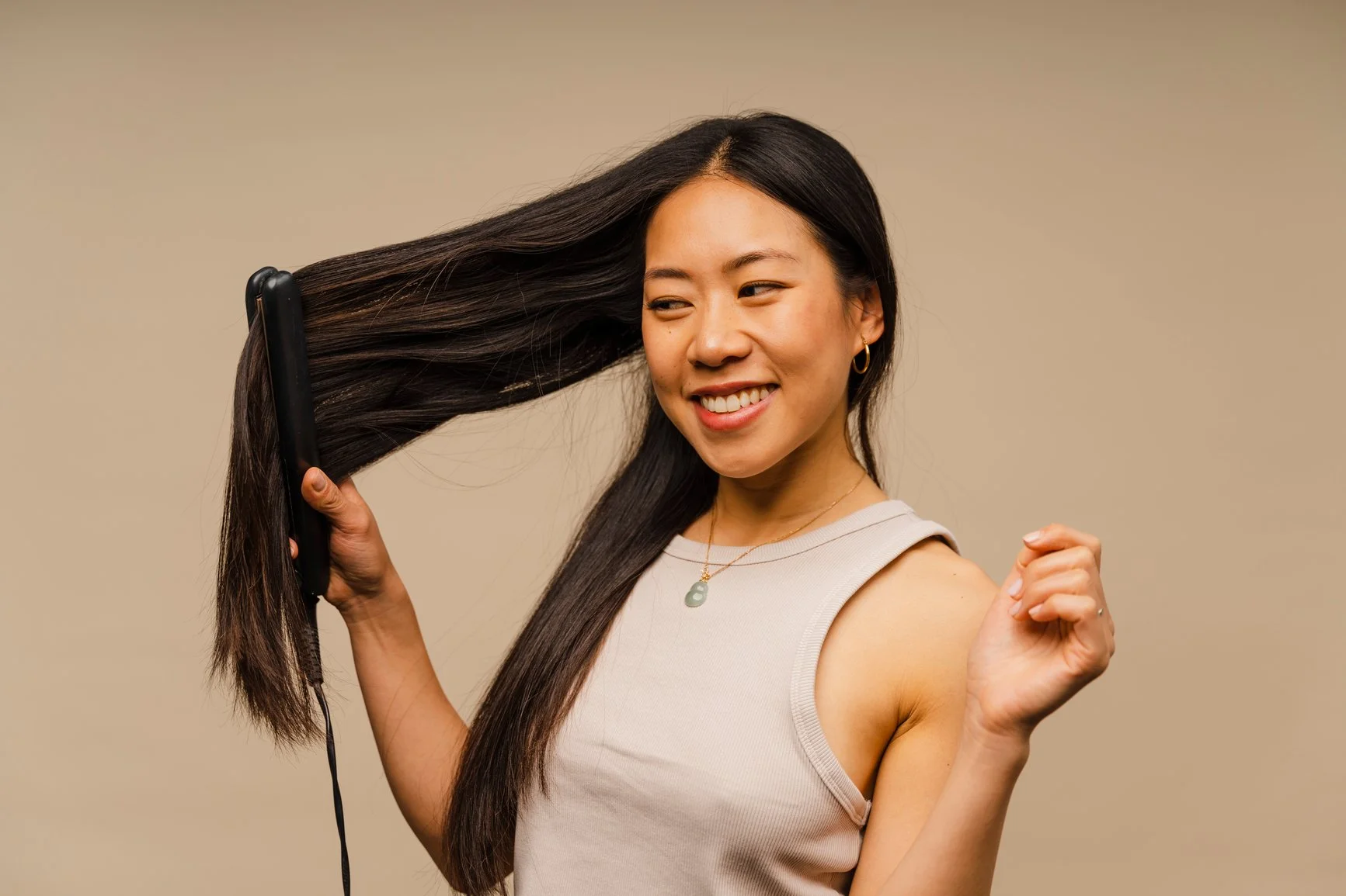 A smiling woman with long, dark brown, sleek hair using a flat iron on her hair.