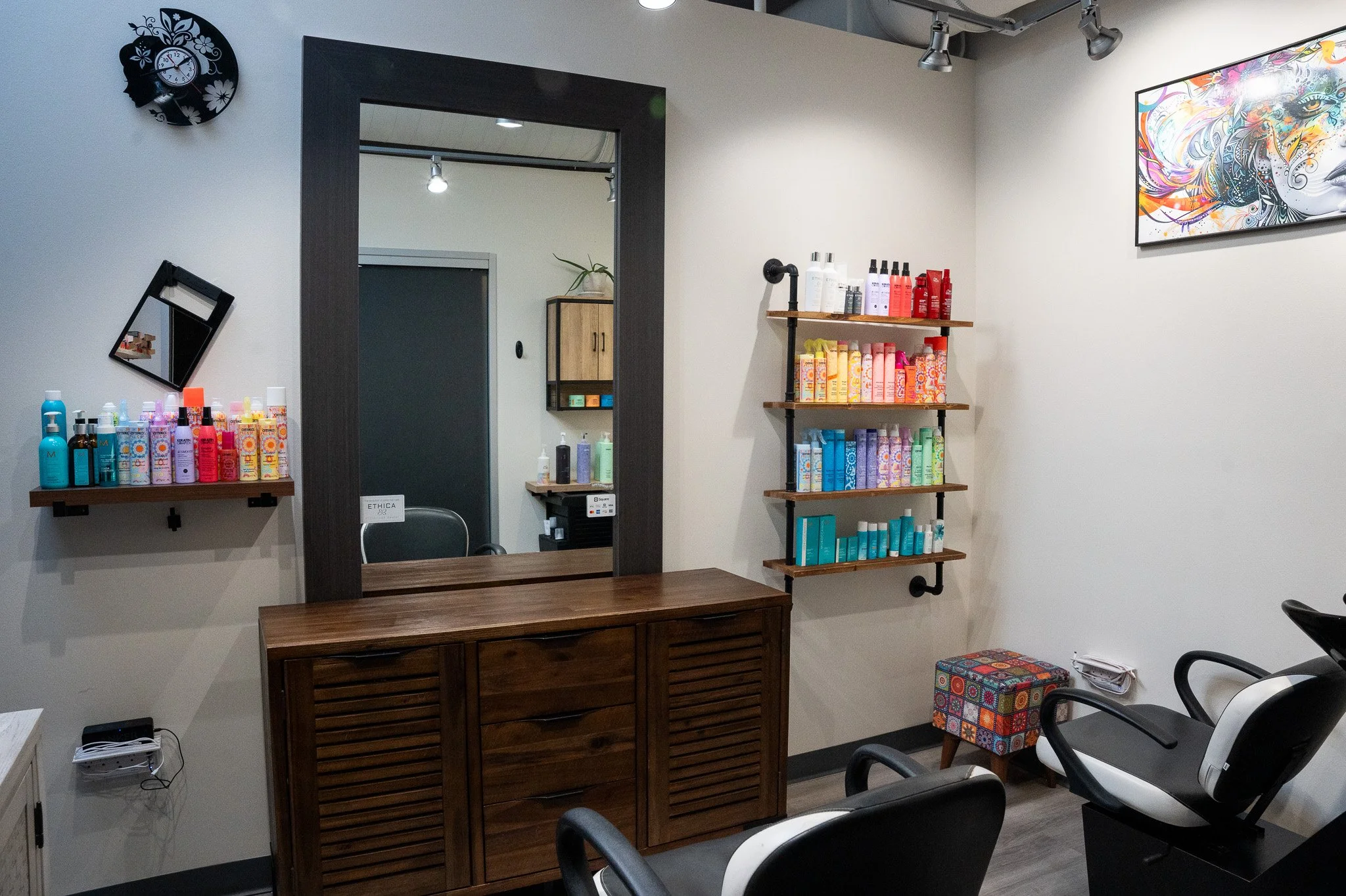 Laura Jayne Hairstylist salon interior with hair products on shelves, a mirror, a wall clock, colorful artwork, and salon chairs.