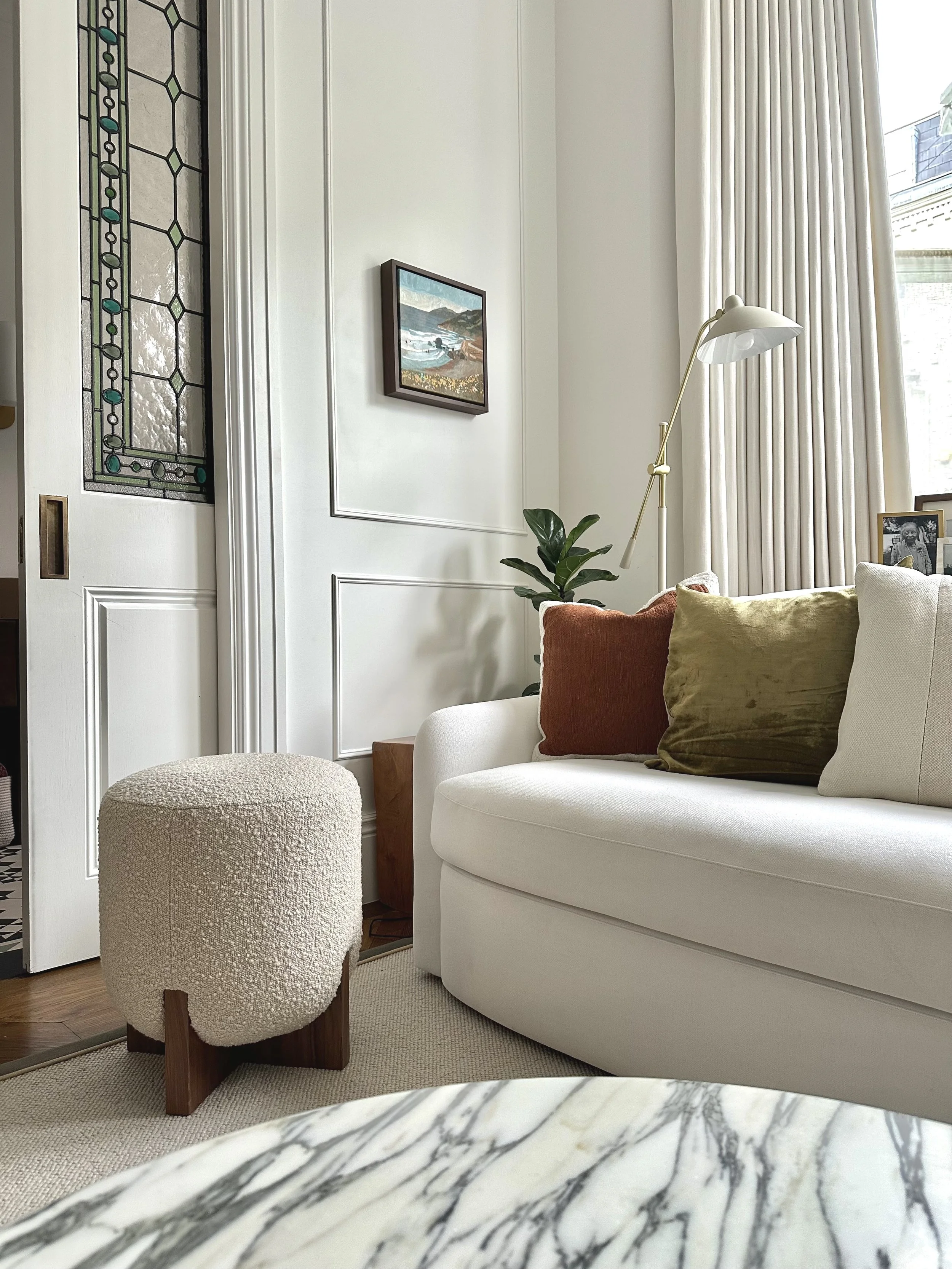 Living room with a white sofa, green and rust-colored pillows, a textured beige ottoman with wooden legs, a tall white floor lamp, a potted plant, a framed photo, and a window with cream-colored curtains.