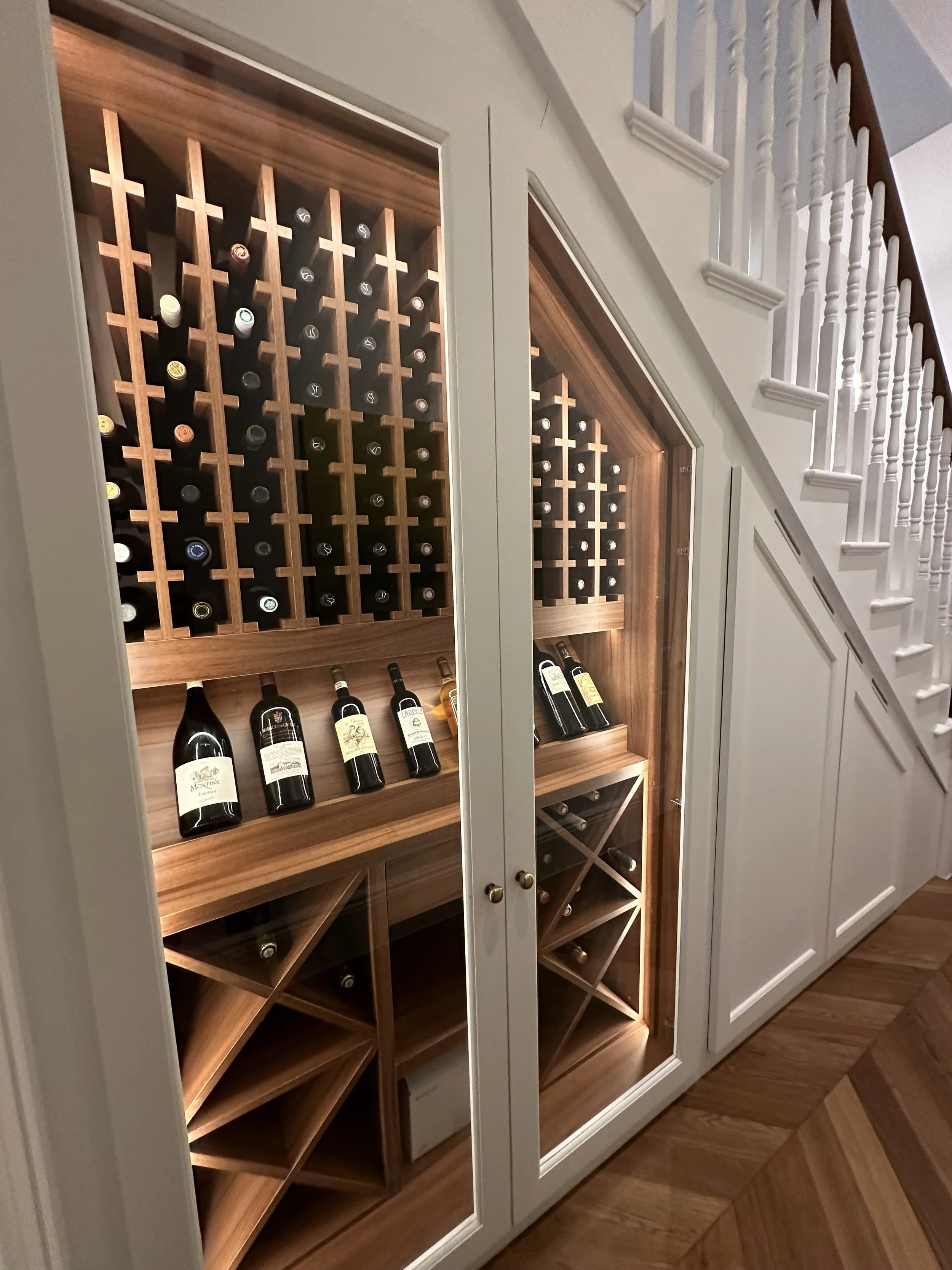 Wine storage built into a white cabinet under a staircase, with bottles stored horizontally and on racks for display.