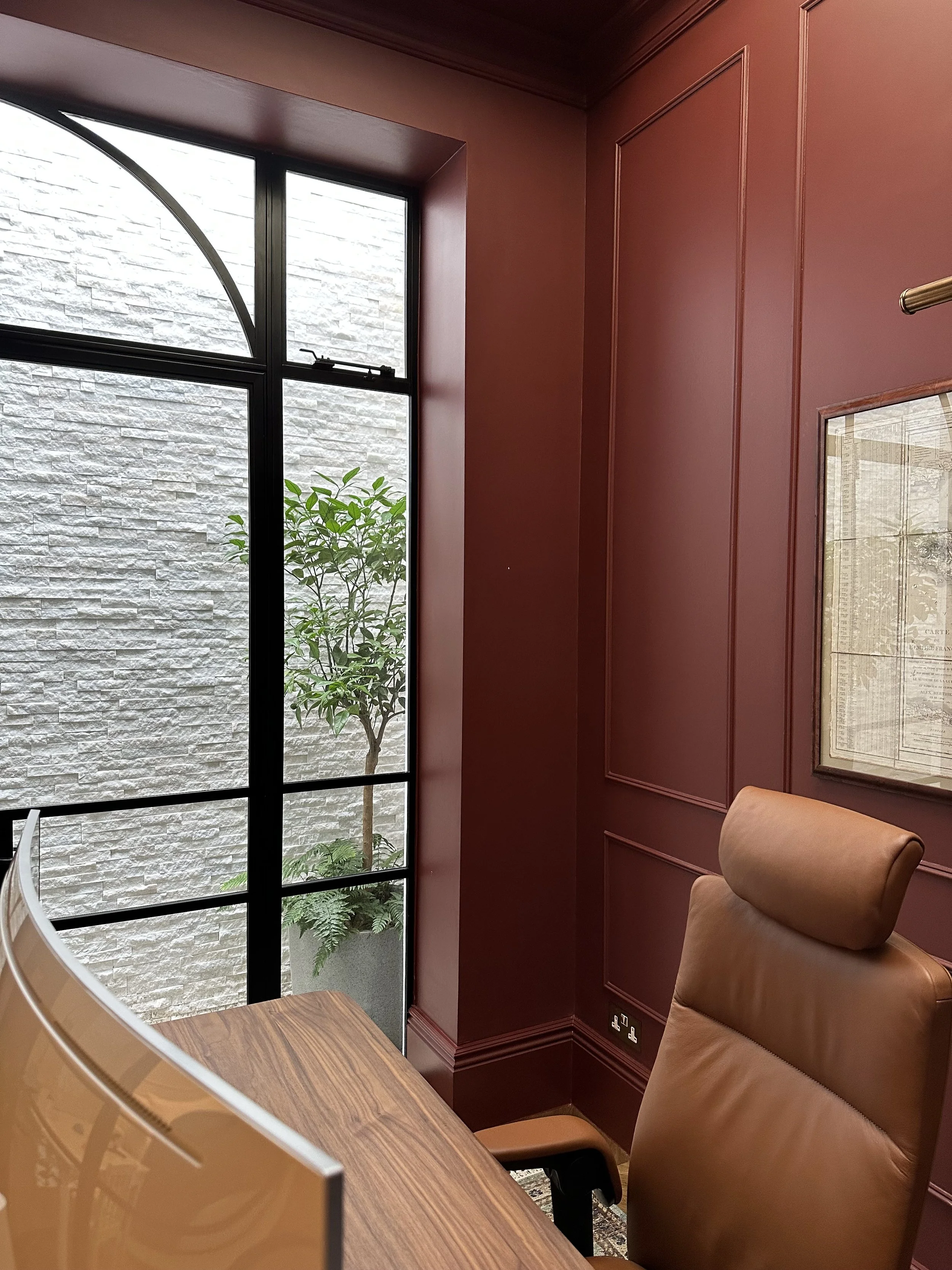Office corner with a leather chair, a wooden desk, red paneled walls, and a window showing a green plant outside.