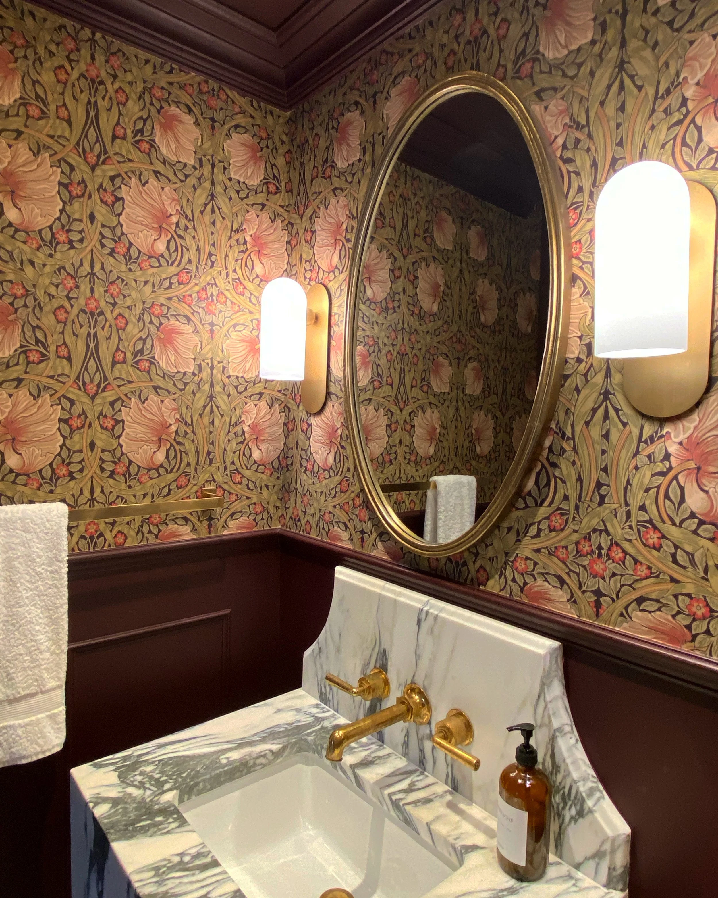 A bathroom with floral wallpaper, an oval mirror with a gold frame, two wall-mounted light fixtures, a marble sink with gold fixtures, and a hand soap bottle.