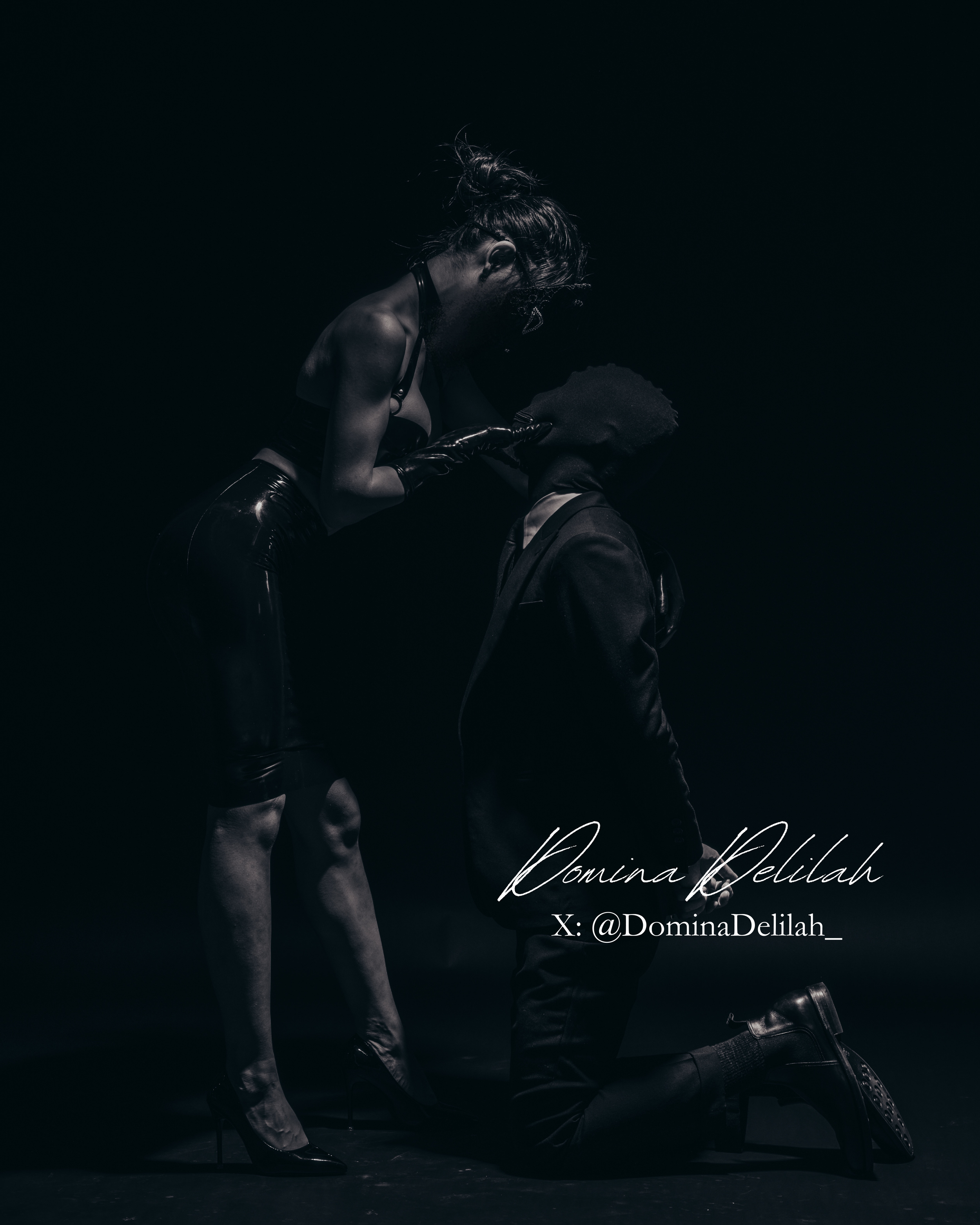 A woman with a bun hairstyle wearing latex clothing and gloves leans over a kneeling person with a box mask and formal wear, in a dark, moody setting.