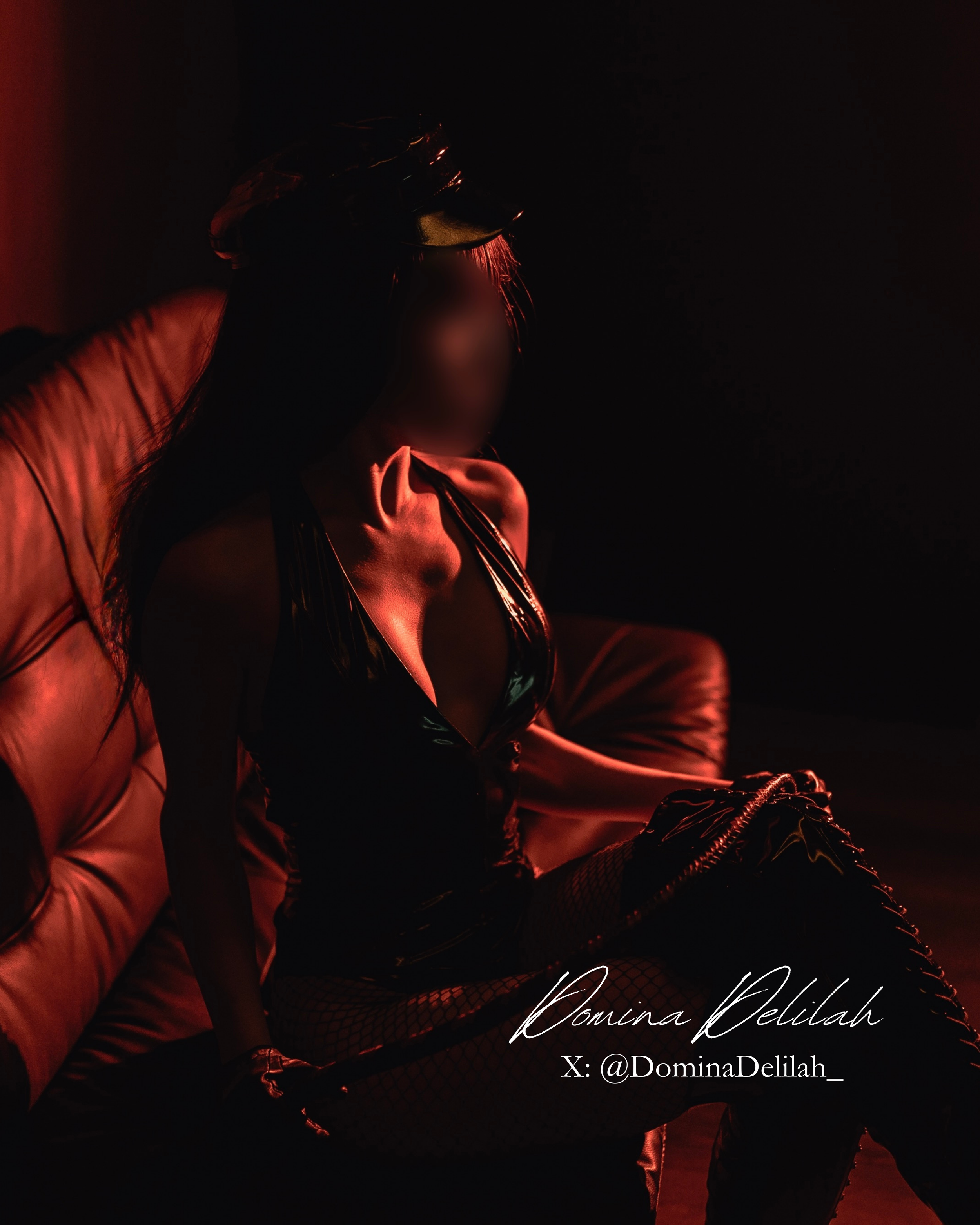 A woman sitting on a leather couch in dim red lighting, wearing a black shiny dress, fishnet stockings, and a hat, with her face blurred. The text overlays read "Domina Delilah" and "X: @DominaDelilah_".