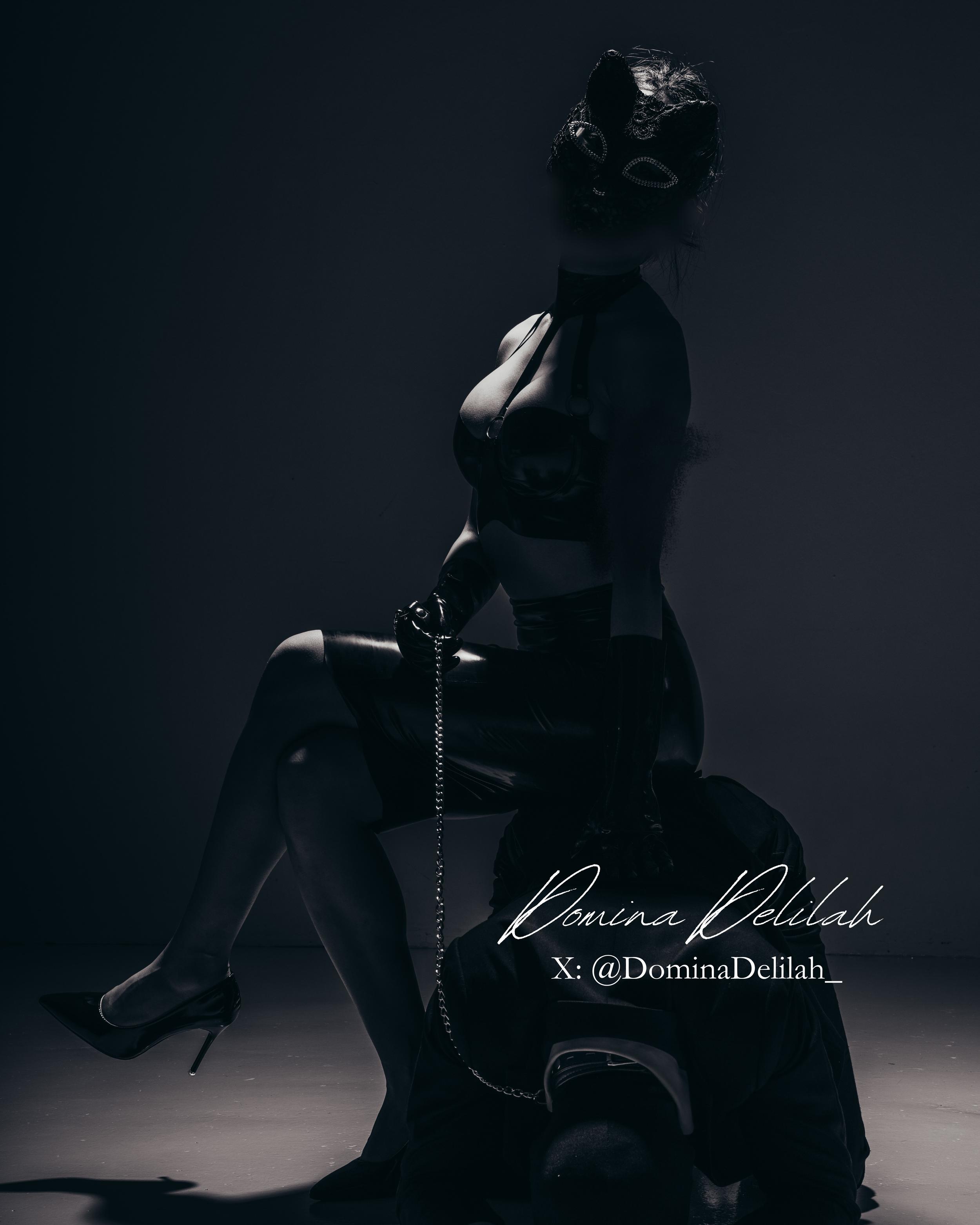 A woman wearing a mask and black leather outfit, sitting on a lead, with dramatic lighting and a shadowy background.