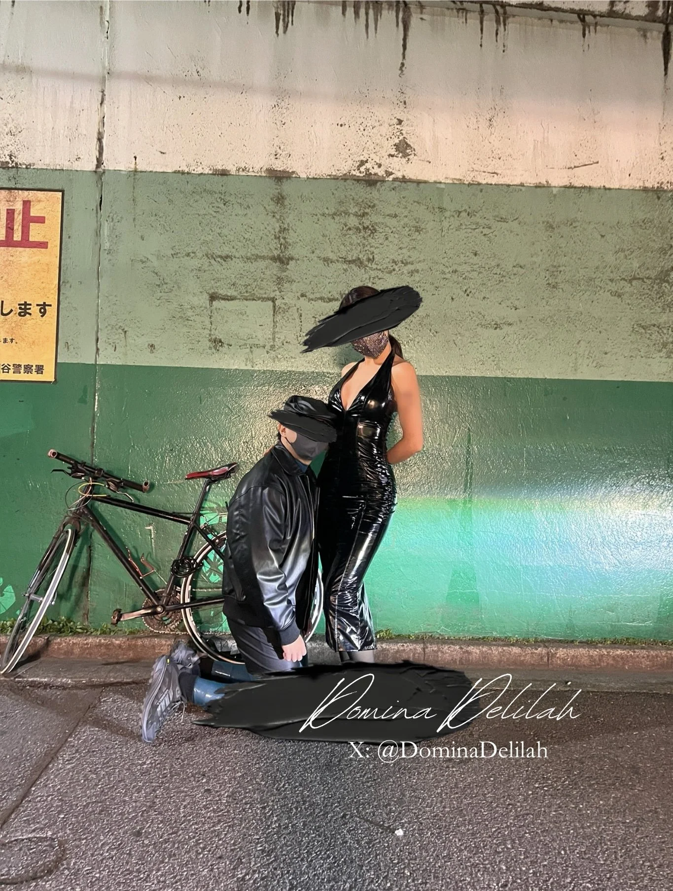 A person dressed in black latex standing on a street, with another person kneeling in front of them, both wearing black masks. There is a bicycle to the left, and the background features a green and beige wall. The image has the text 'Romina Delilah' and '@DominaDelilah' written at the bottom right.