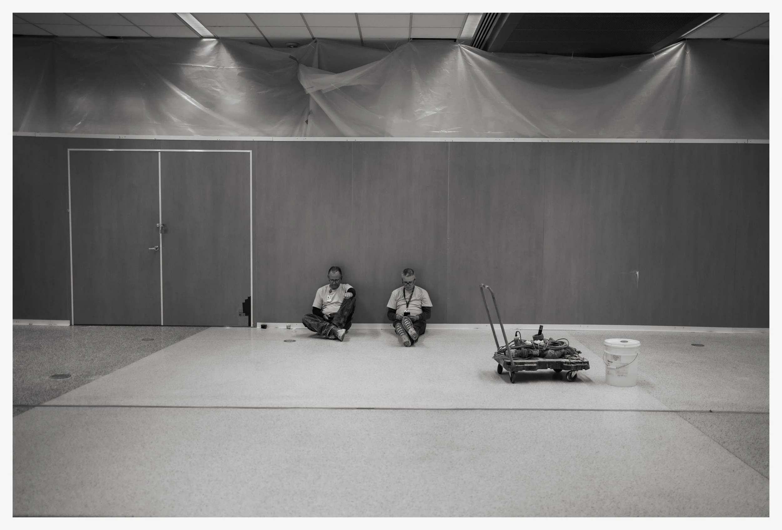Two workers sitting on the floor against a plain wall, with cleaning equipment including a flat mop on a trolley and a bucket nearby.