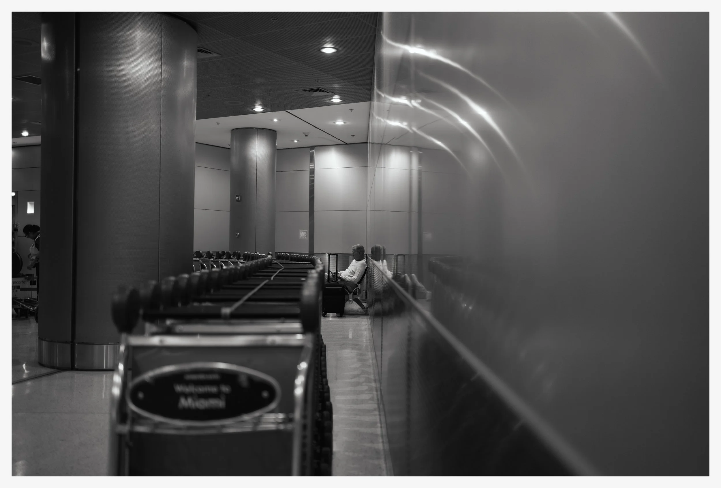 Black and white photo of an airport waiting area with metal pillars and a row of luggage carts. A man is sitting on a bench facing away, and a woman in the background is working with a cart.