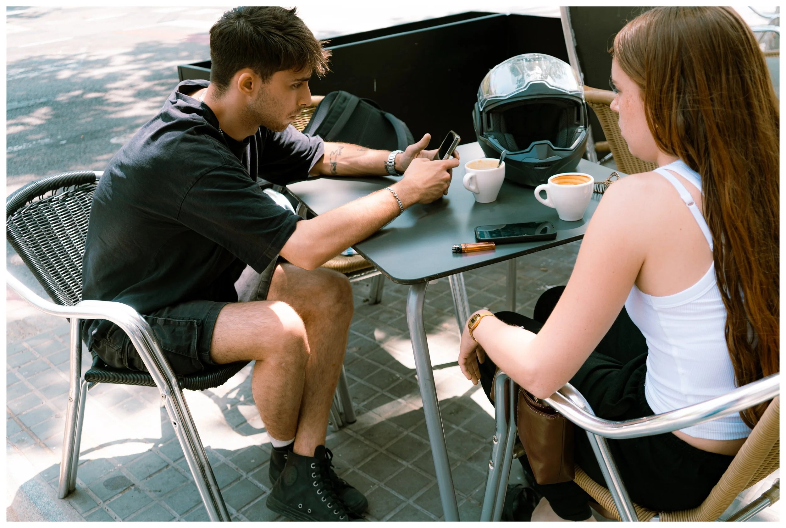 A young man with short brown hair, wearing a black T-shirt and shorts, sitting at an outdoor table, looking at his phone. A young woman with long red hair, wearing a white tank top and black pants, sitting across from him. The table has two cups of c