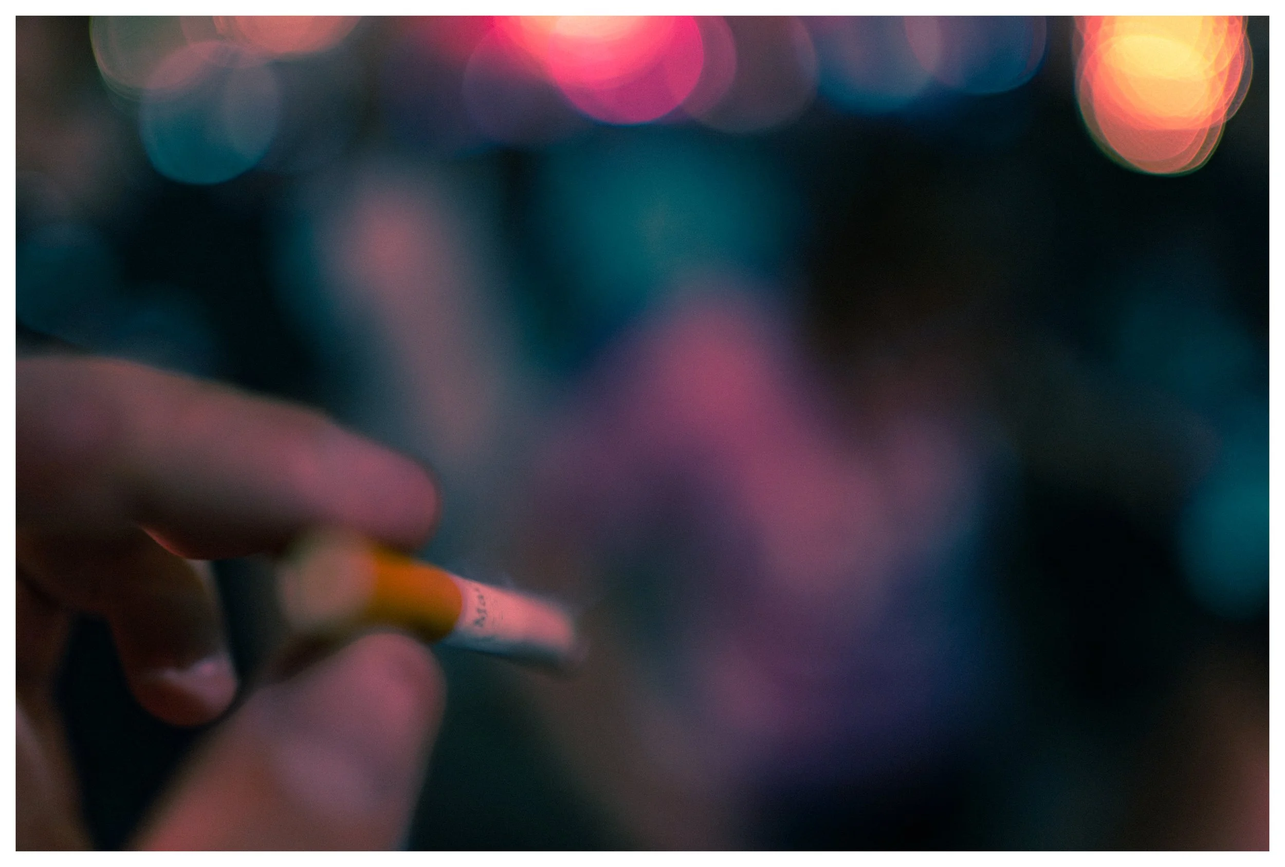 Close-up of a person's hand holding a cigarette, with colorful bokeh lights in the background.
