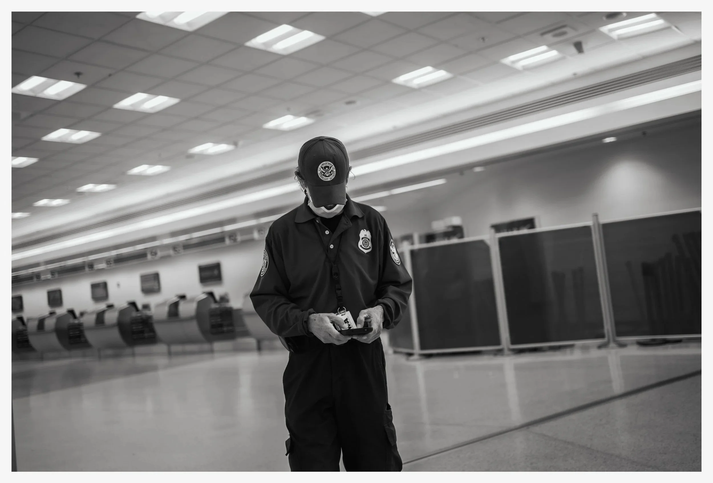 A subway or train station attendant wearing a dark uniform, a cap with an emblem, and a face mask, standing alone in an empty station area with ticket kiosks and monitors in the background.