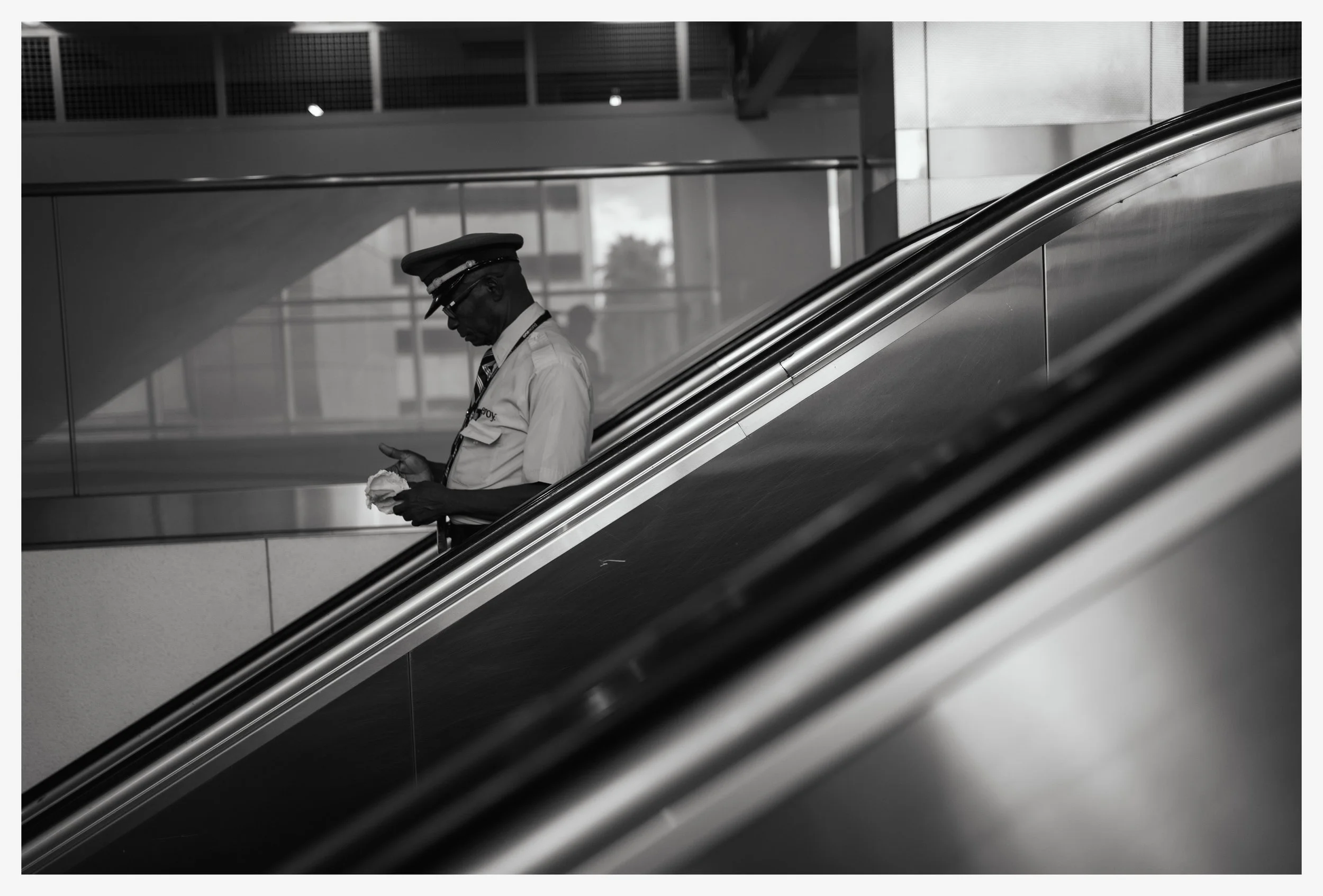 A black-and-white photo of a security guard or officer leaning on an escalator railing while looking at his phone, wearing a uniform, cap, and glasses, in a modern indoor public space.