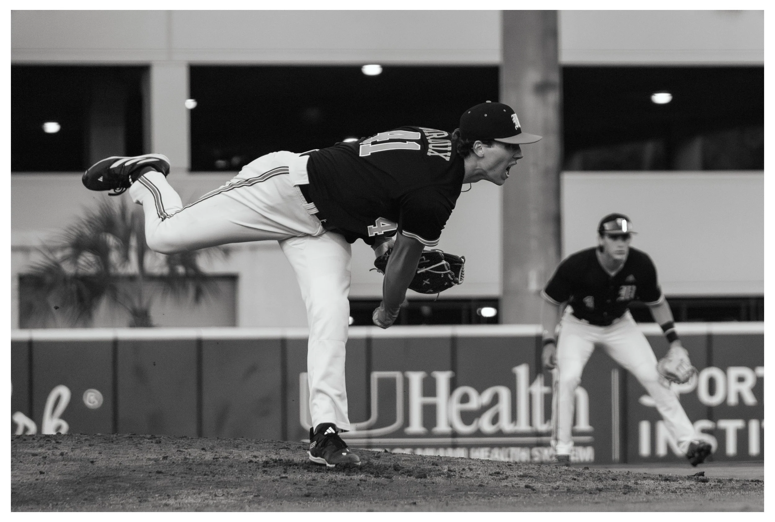 A baseball player in a black jersey and white pants is leaning forward, mid-pitch on the mound during a game. Another player in similar uniform is in the background, ready on the field.