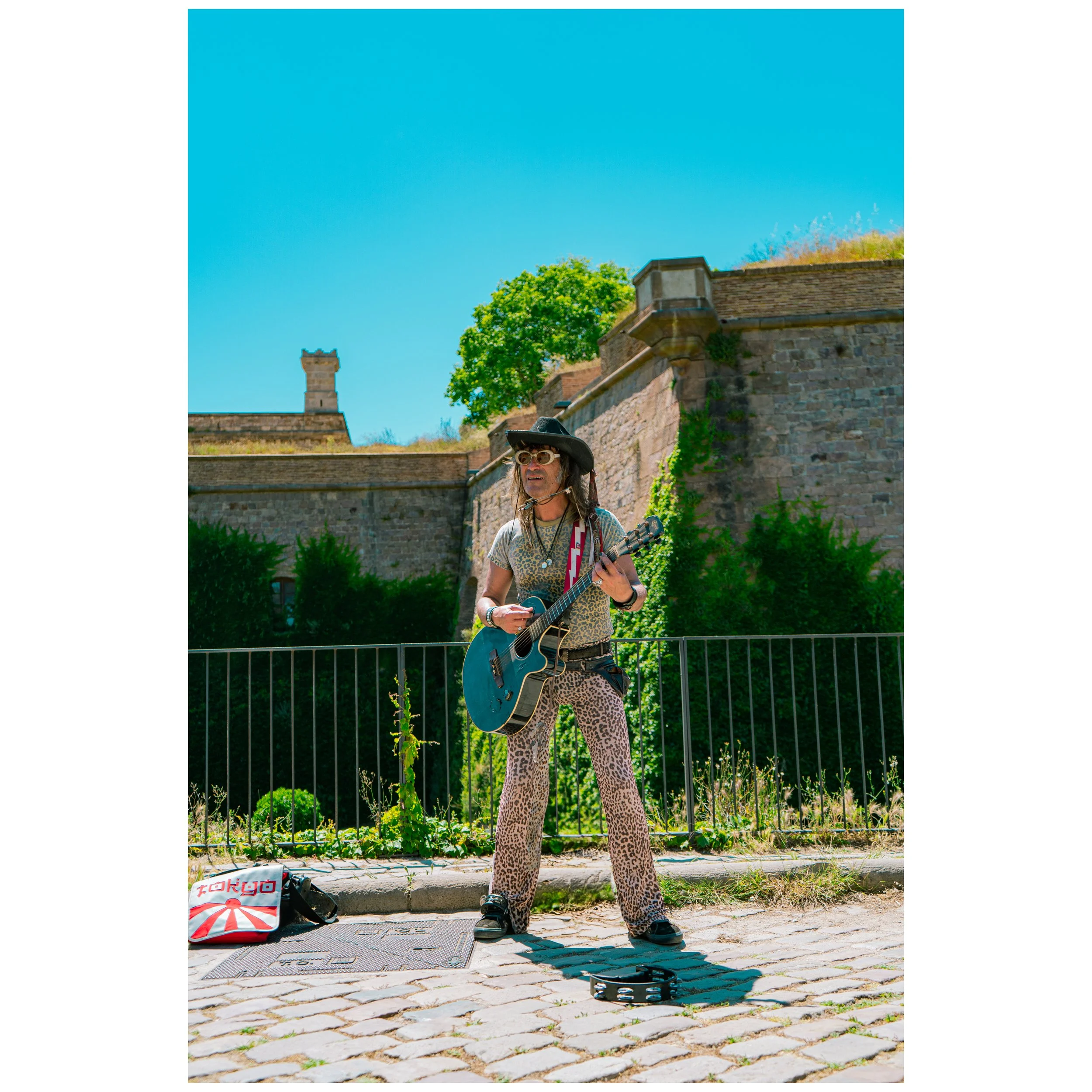 A street performer with long hair, wearing a black hat, sunglasses, leopard print pants, and a patterned shirt, plays a blue acoustic guitar on a cobblestone street under a bright blue sky. A bag with Tokyo written on it and a tambourine are on the ground nearby.
