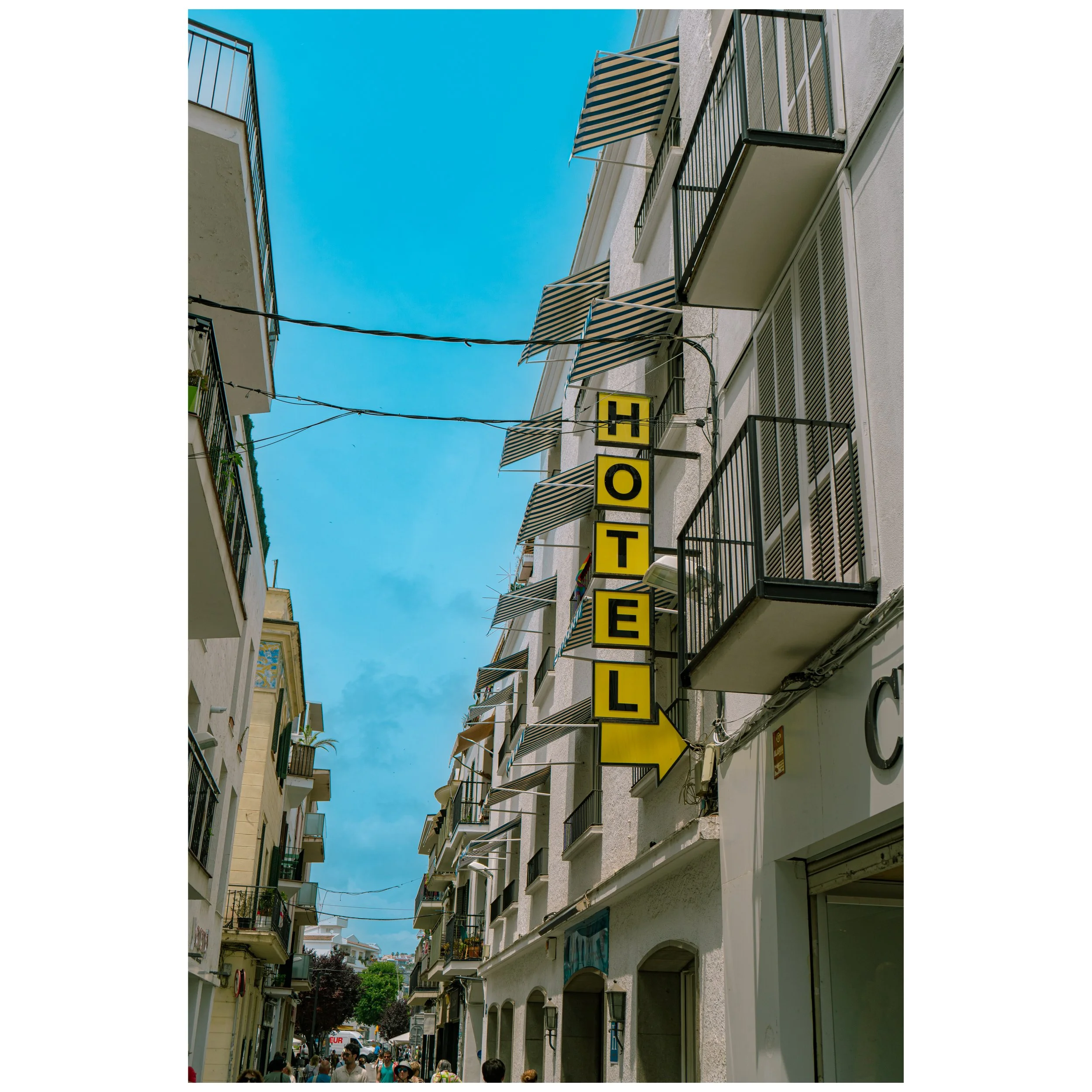 A narrow street lined with multi-story white buildings with balconies. A vertical yellow sign with black letters spelling 'HOTEL' and an arrow pointing to the right is attached to one of the buildings. The sky is bright blue with some clouds, and there are people walking on the street.