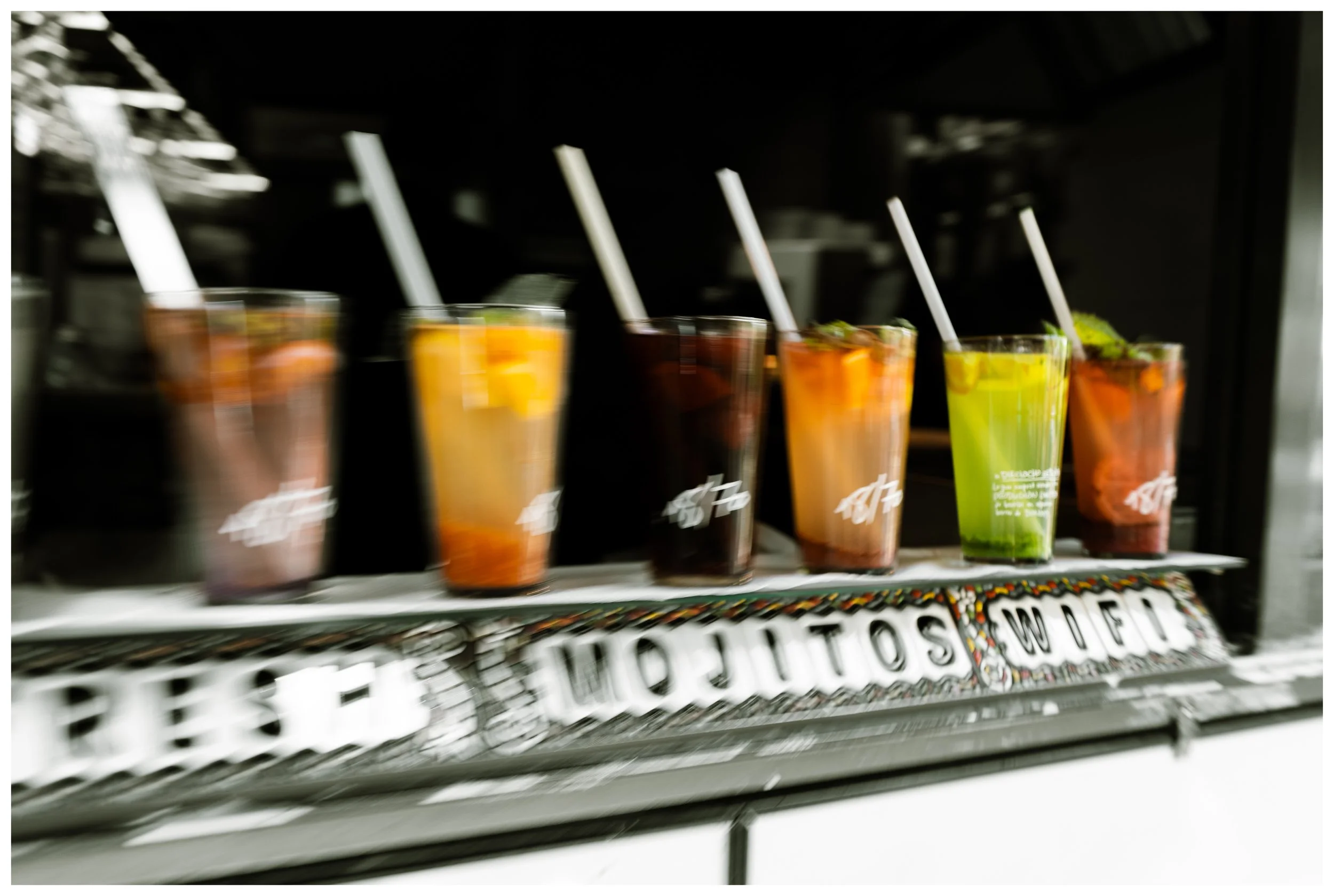 Six colorful drinks in clear cups with straws on a bar counter, with a blurred background.