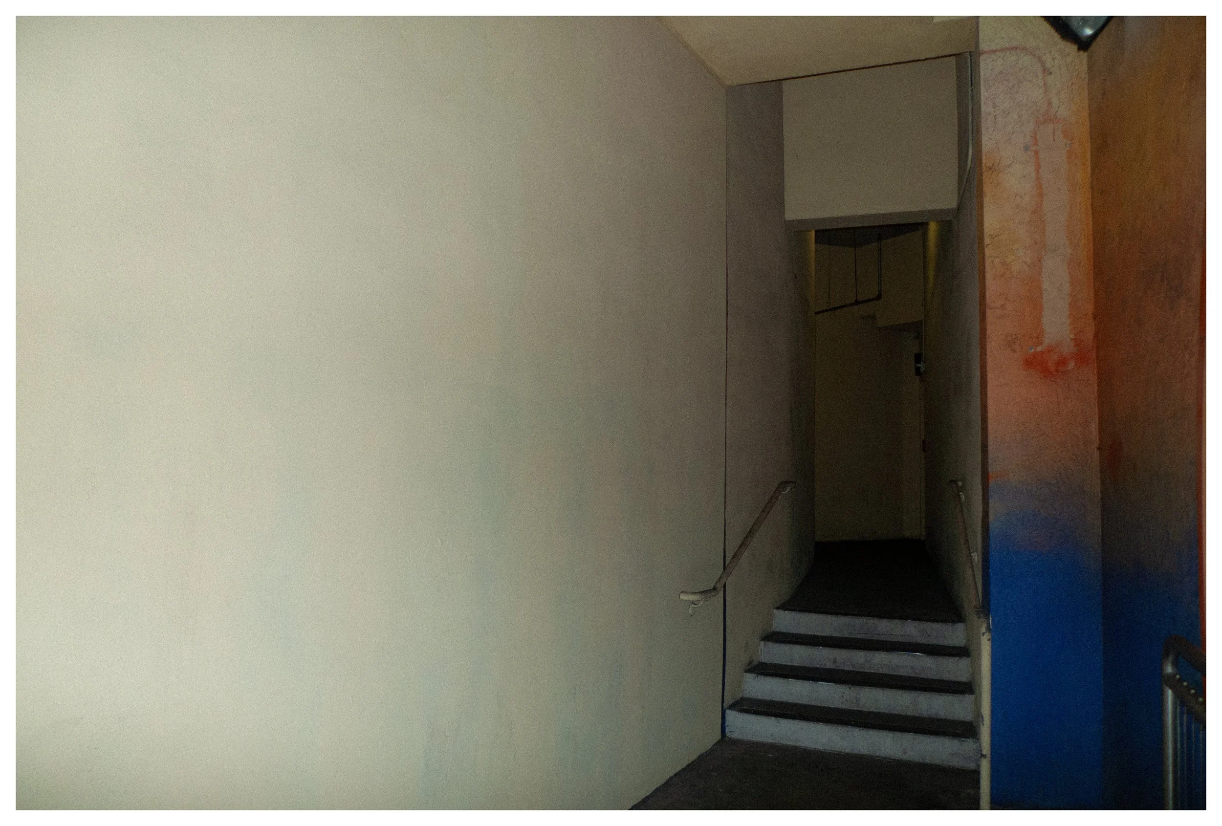 A stairway with few steps leading upward, next to a plain, light-colored wall. The staircase has a metal handrail on the left side, and an opening at the top of the stairs leads into a dimly lit corridor. On the right side of the image, there is a wa