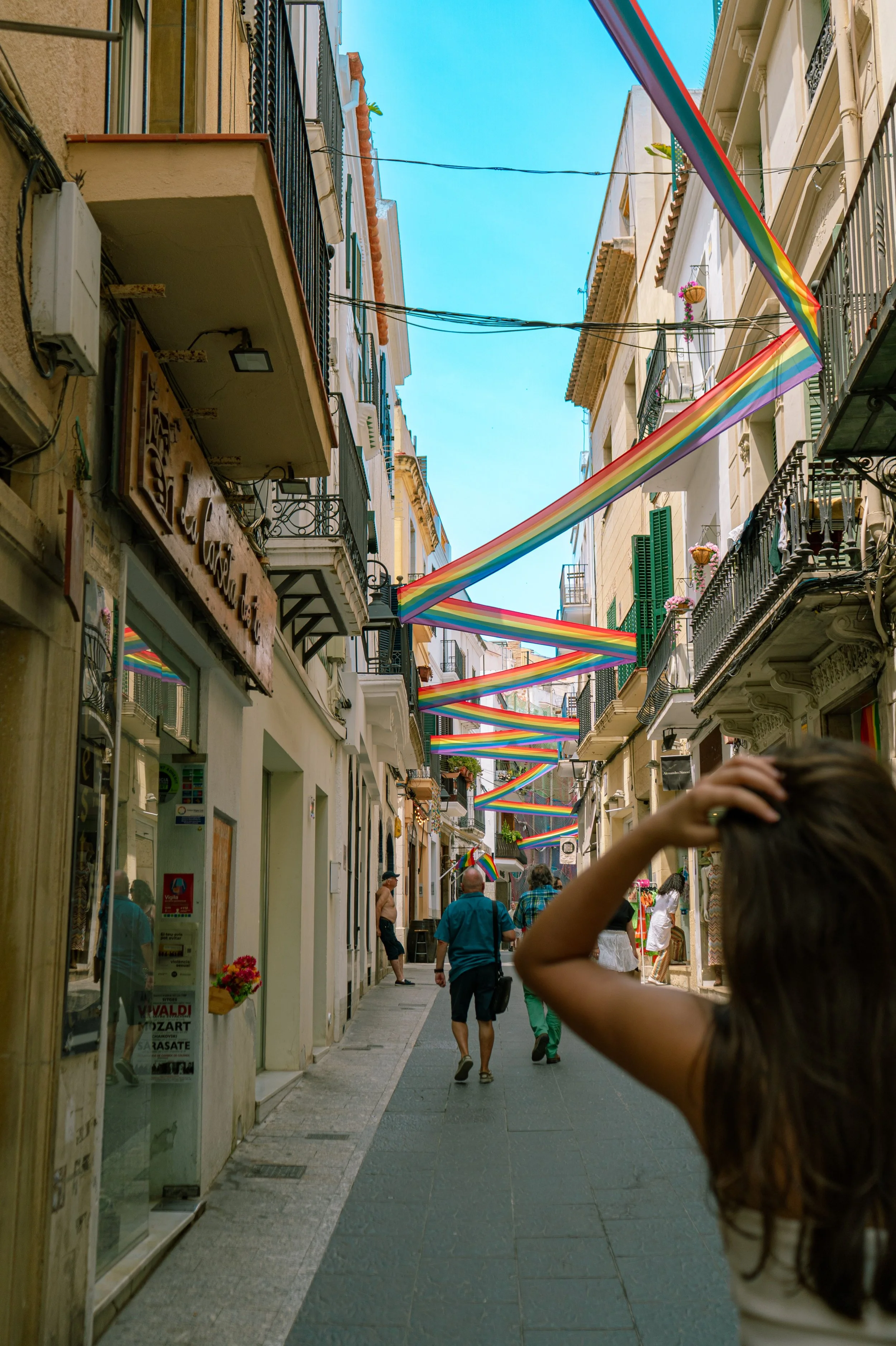 A street with colorful rainbow banners overhead and people walking along the sidewalk.