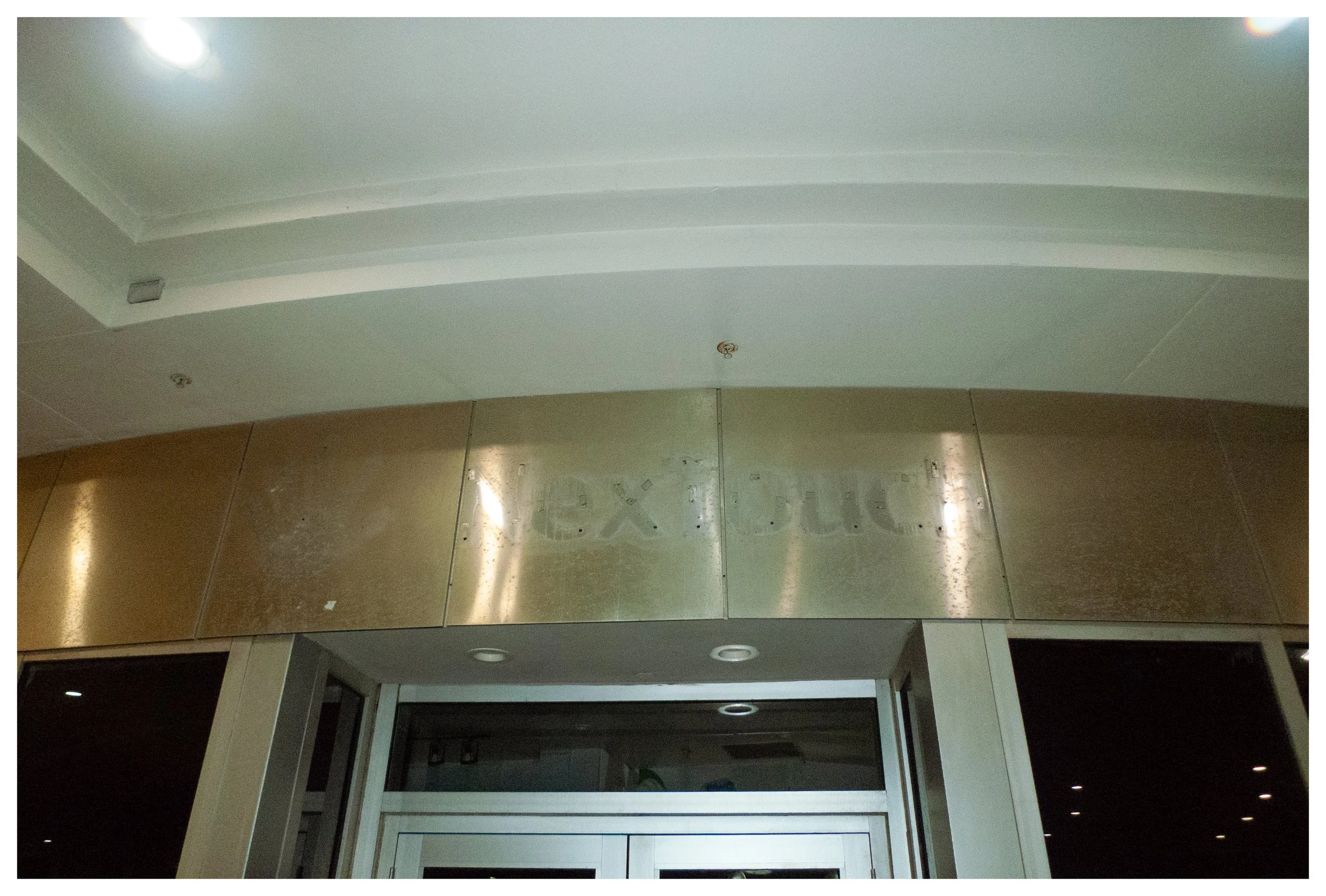 Close-up of a metallic sign that reads 'Lex D.C.' above entrance doors, with part of the ceiling and lighting visible.