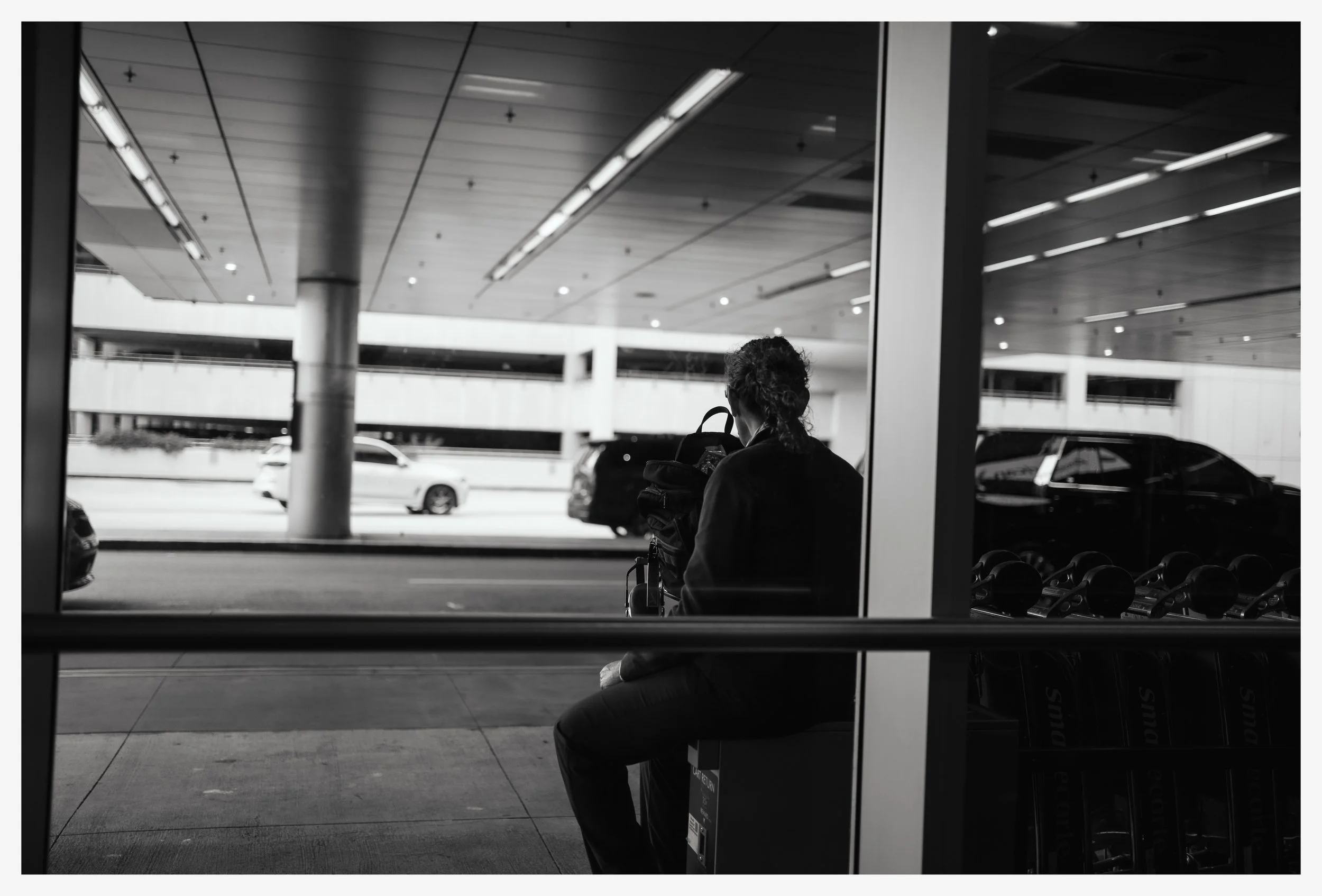 A woman sitting outside at an airport terminal, looking at her backpack. The image is in black and white.