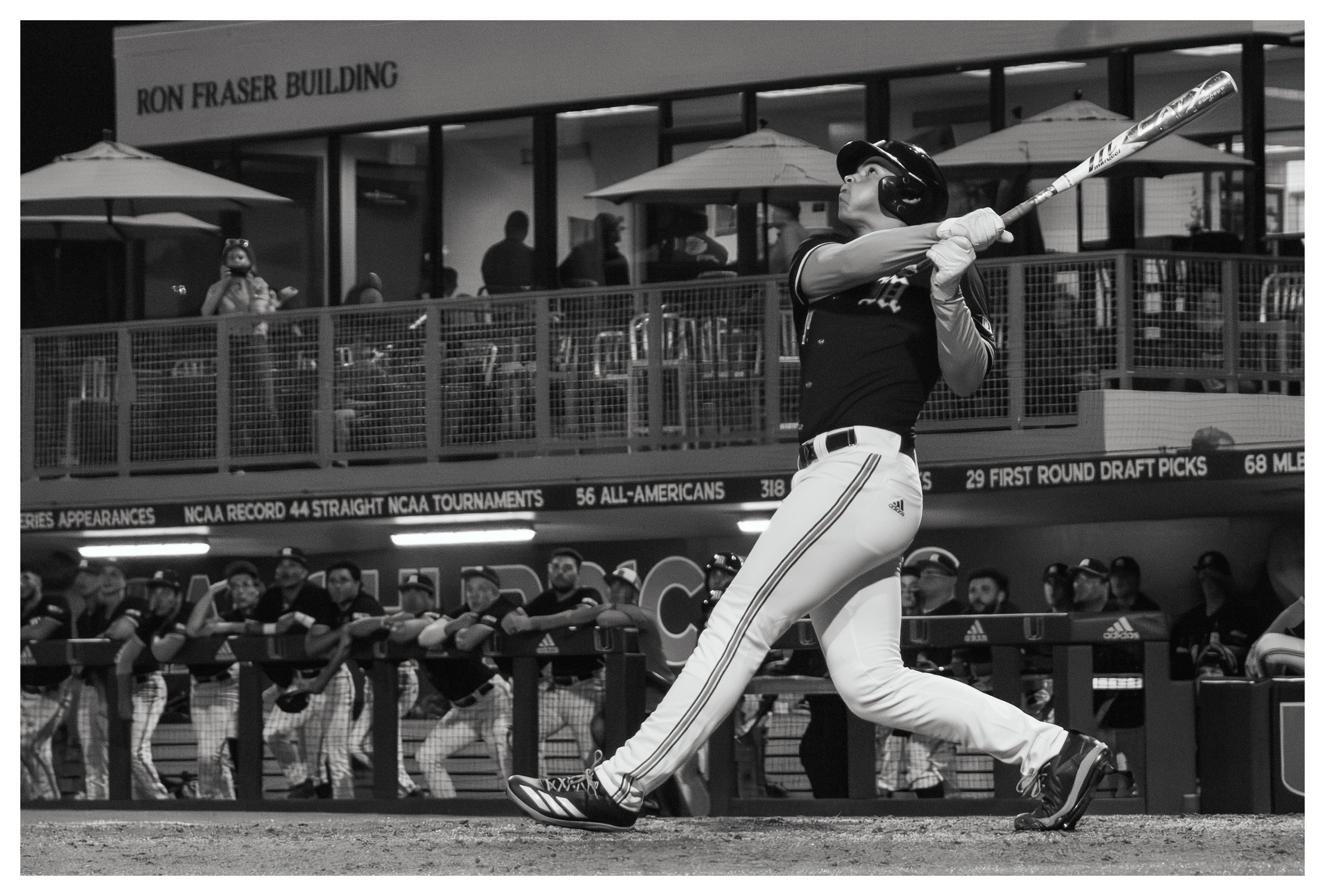 A baseball player swinging a bat during a game, with teammates in the dugout and spectators in the background.