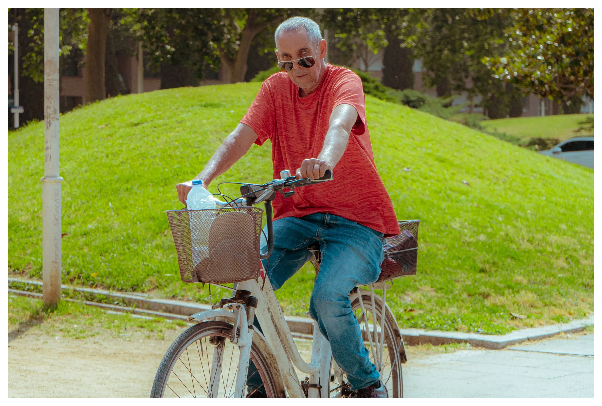 An older man wearing sunglasses and a red t-shirt, riding a bicycle on a sunny day in a park with green grass and trees in the background.