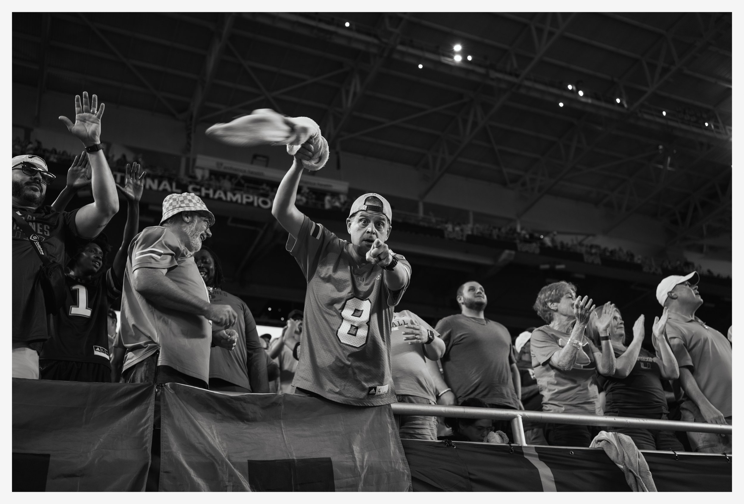 Group of football fans standing at stadium, some clapping and cheering, one person holding a hat, black and white photo.
