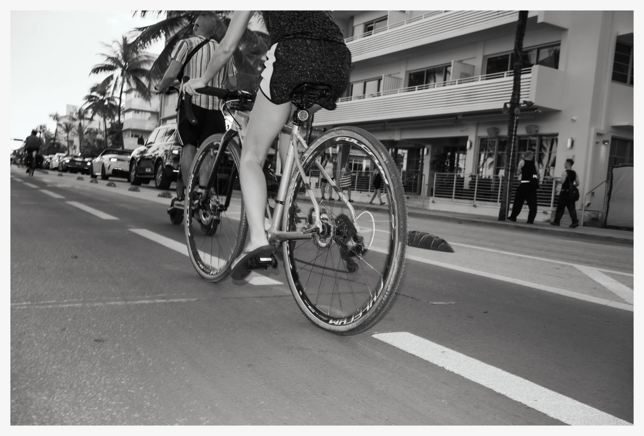 A cyclist riding a bike on a city street with parked cars and pedestrians in the background, captured in black and white.