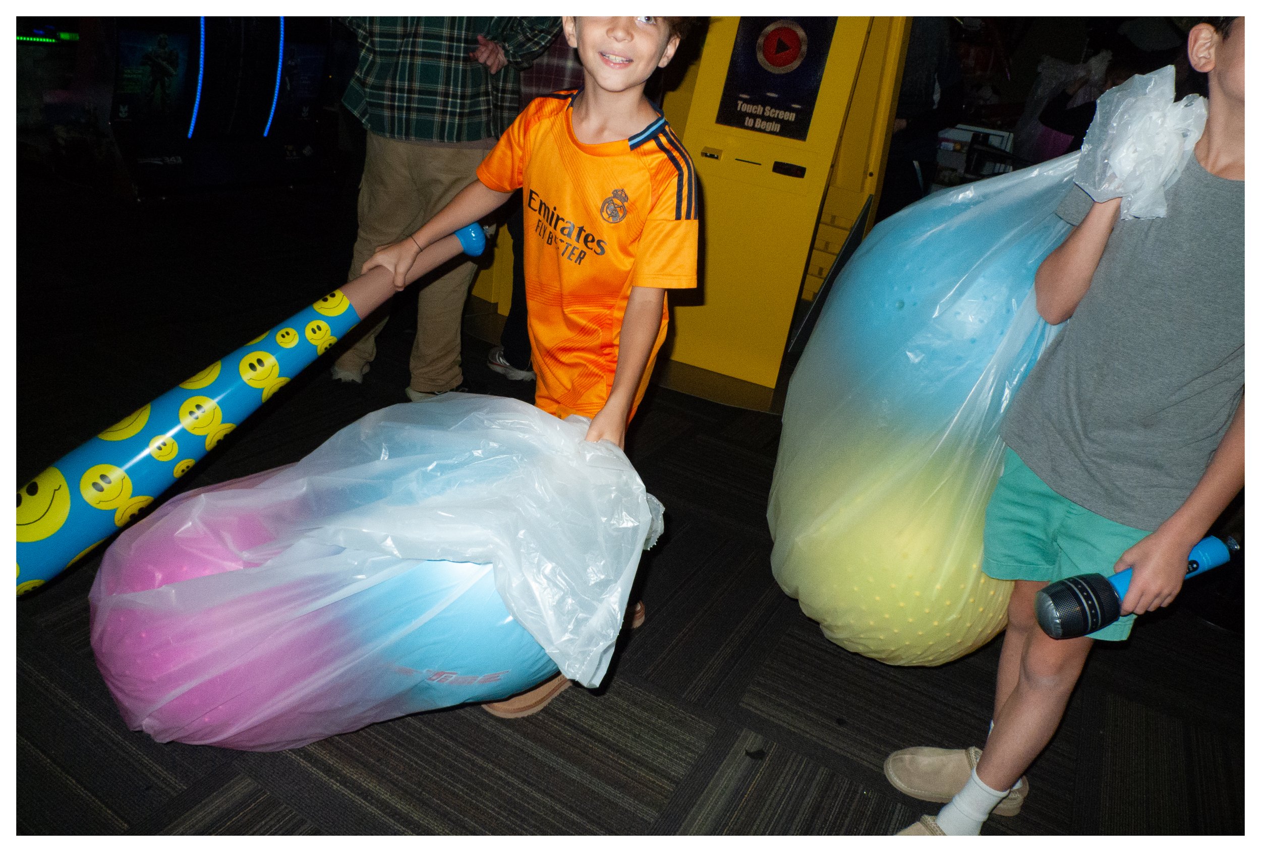 Two children holding large inflatable balls wrapped in plastic and decorated with smiley face wrapping paper, standing in an arcade.