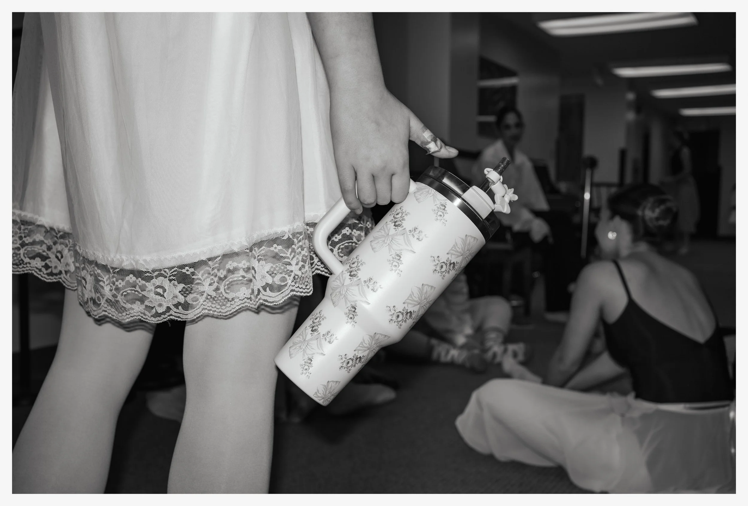 A person is holding a floral-patterned water pitcher behind their back, while sitting on the floor of a room with other seated individuals in the background.