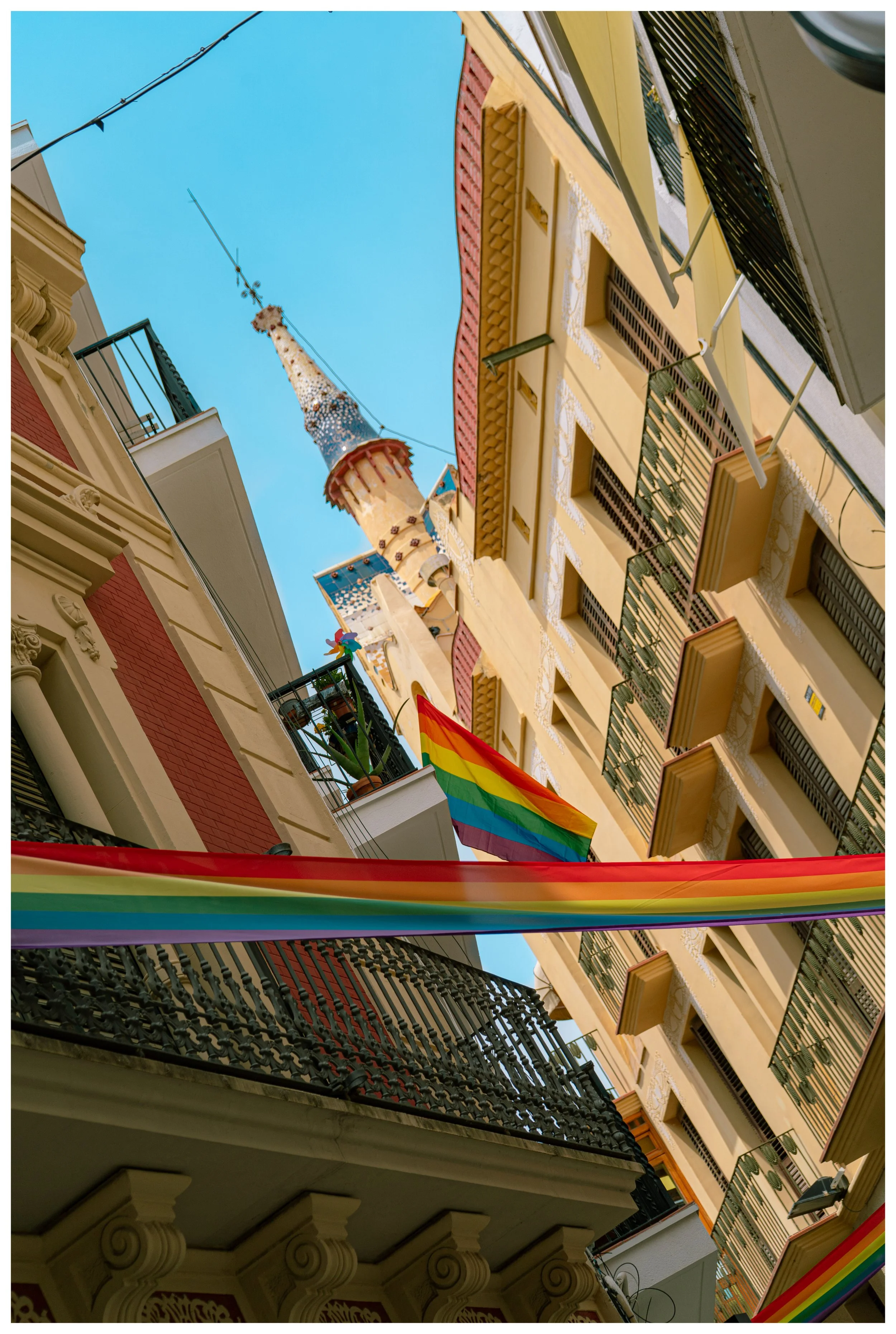 View of colorful buildings with balconies, a rainbow flag, and a rainbow fabric, with a church steeple and a clear blue sky in the background.