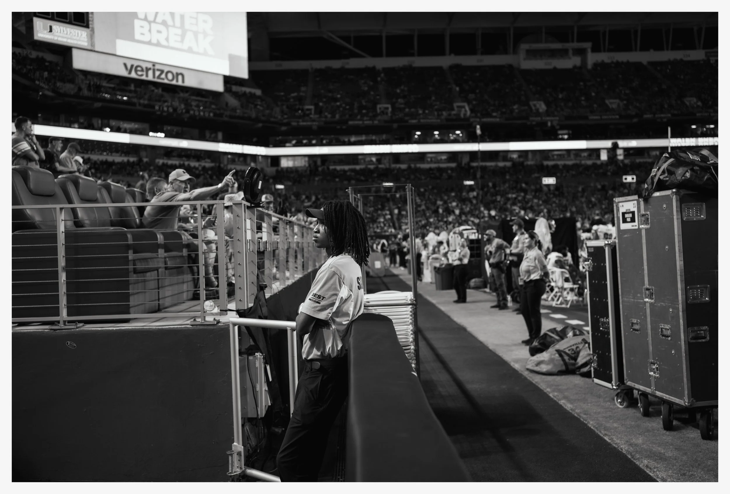 A woman with dreadlocks leaning against a barrier on the sidelines of an indoor stadium, watching a sporting event. Audience members and officials are visible, with some standing and others sitting, and sports equipment and storage cases are nearby.