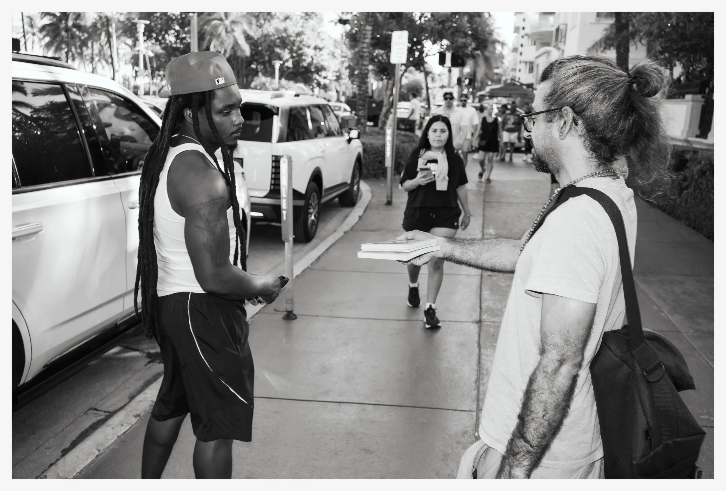 A man with long dreadlocks wearing a cap, tank top, and shorts receives a flyer from another man with long hair tied back, glasses, and a backpack on a city sidewalk. A woman with dark hair and shorts walks behind, looking at her phone, with trees an