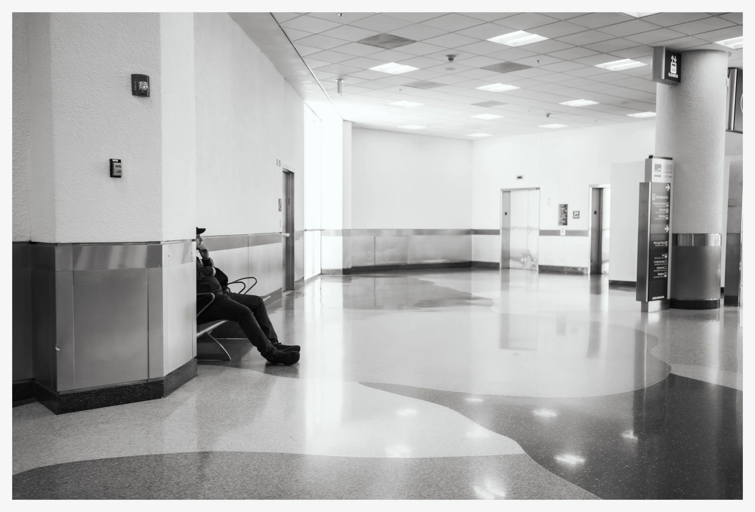 An empty airport terminal with a man sitting alone on a bench on the left side of the image. The floor has a shiny, patterned surface with a curving design. In the background, there are elevators and a sign with directions.